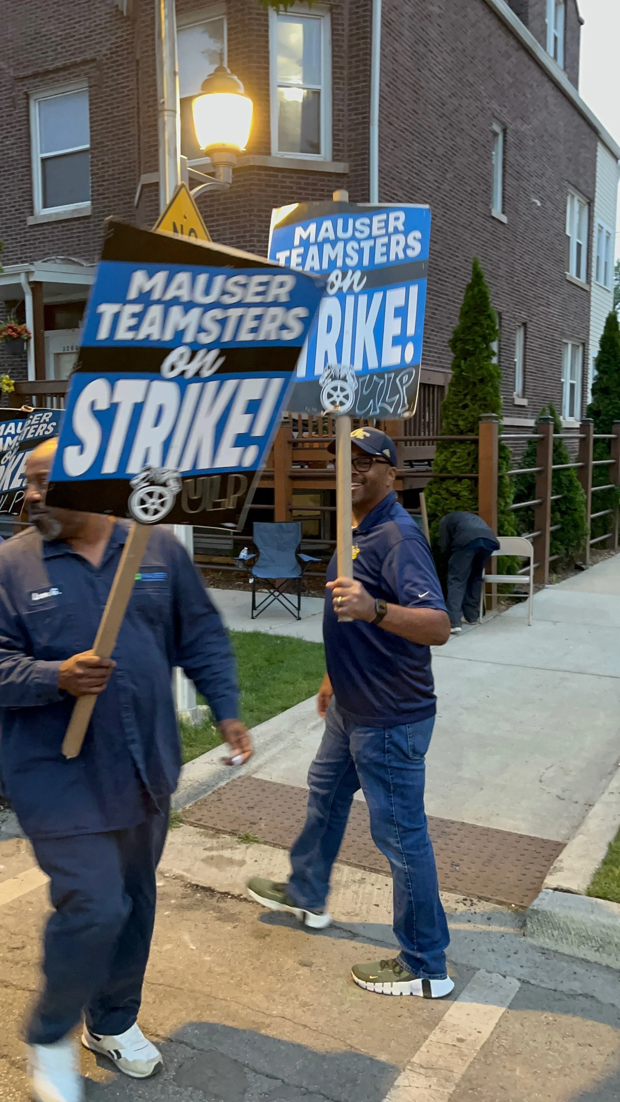 Thomas Gary on the picket line holding protest signs that read 'Mauser Teamsters on Strike!'. They are standing on a sidewalk near a residential area with a brick building and a street lamp in the background.