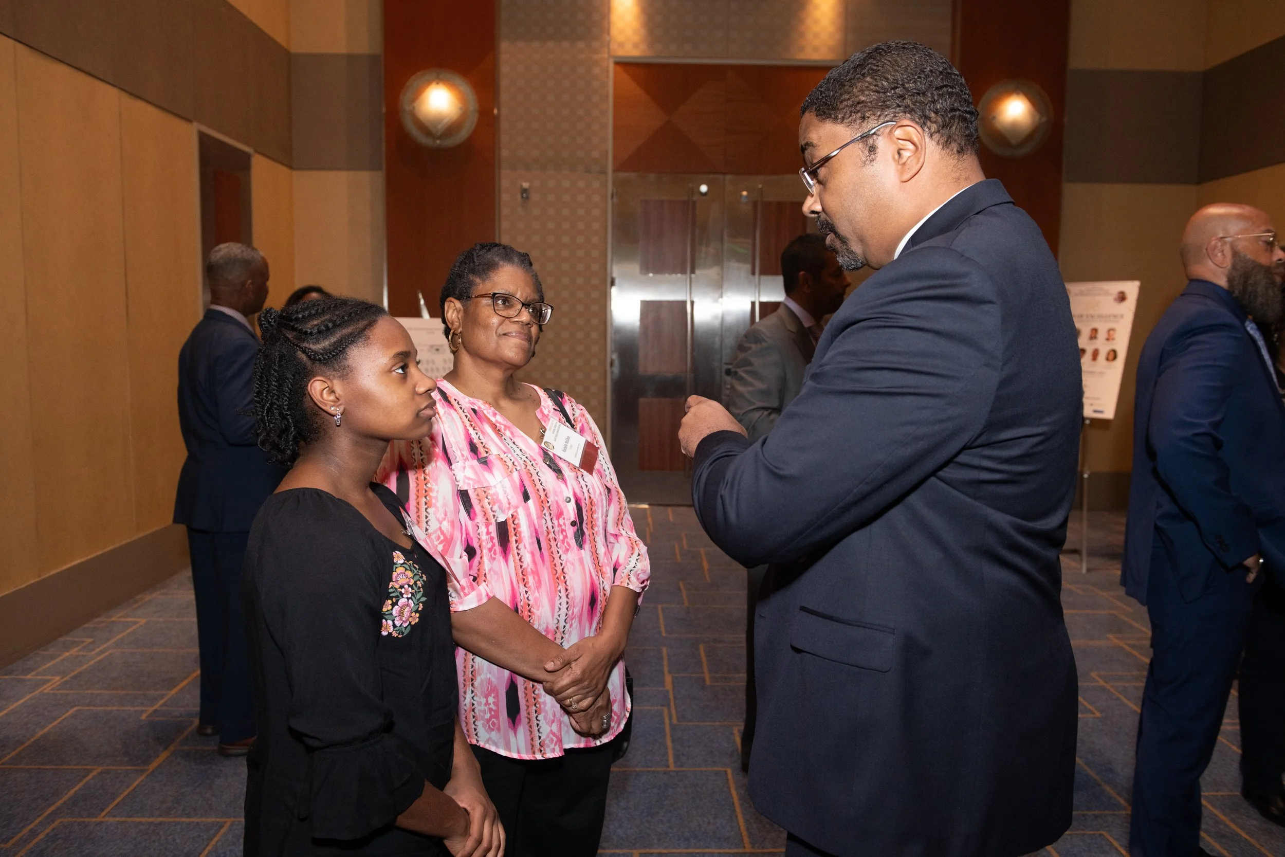 Thomas Gary having a serious conversation at a professional event, with two women and one man, all dressed in business attire, standing in a conference room with wood-paneled walls and carpeting.