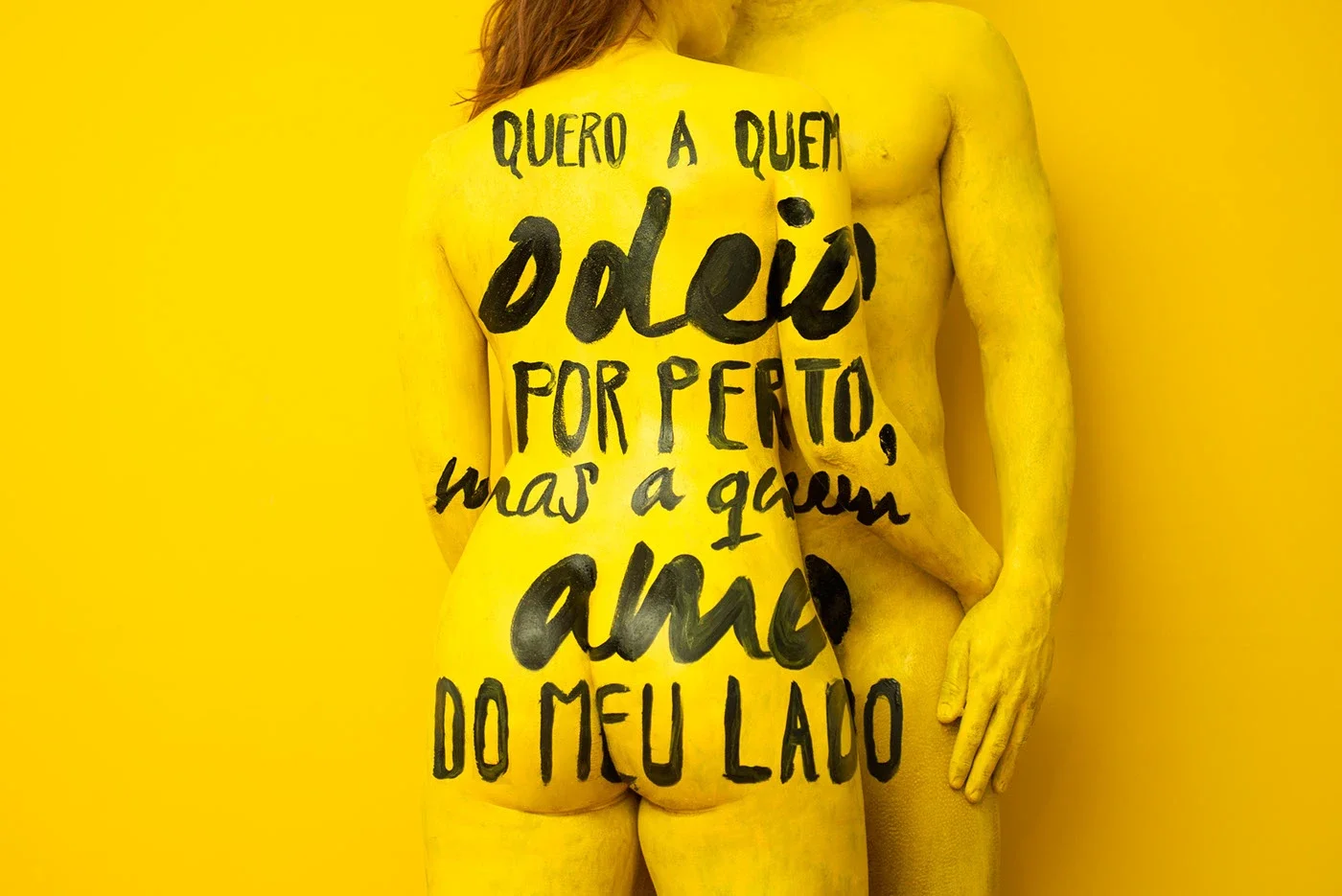 Pessoa de costas pintada de amarelo, com a frase escrita em preto no corpo, contra um fundo amarelo.