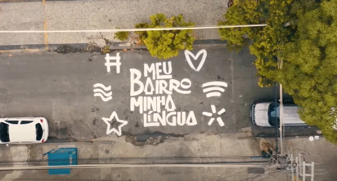Arte de rua com letras brancas na rua dizendo 'Meu bairro minha língua' e desenhos de coração, estrela, ondas e outros símbolos.