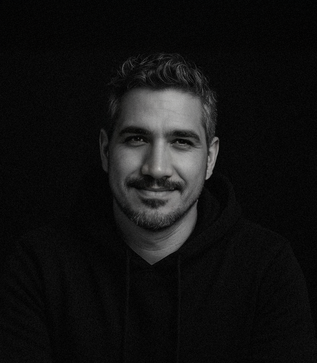 Retrato de um homem com cabelo curto, barba por fazer, sorrindo, com fundo preto e iluminação suave.