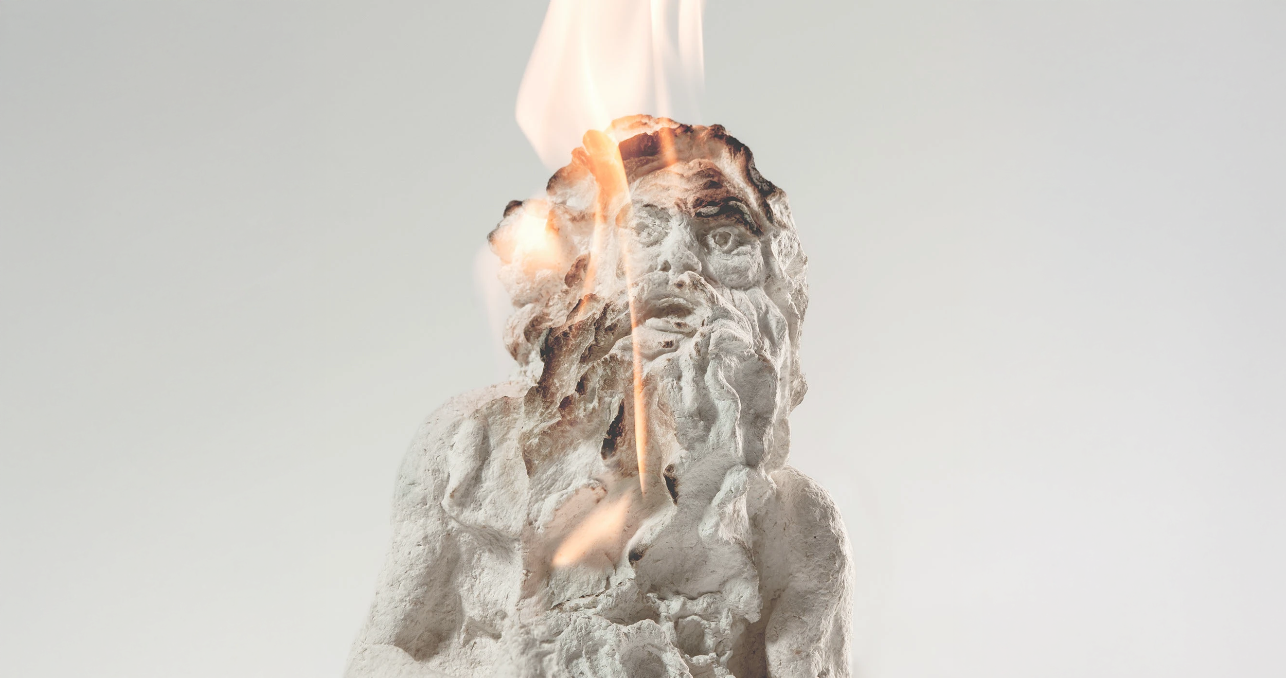 Escultura de um rosto humano com detalhes de uma barba e cabelo, bombardeada por fogo e fumaça, contra um fundo branco.