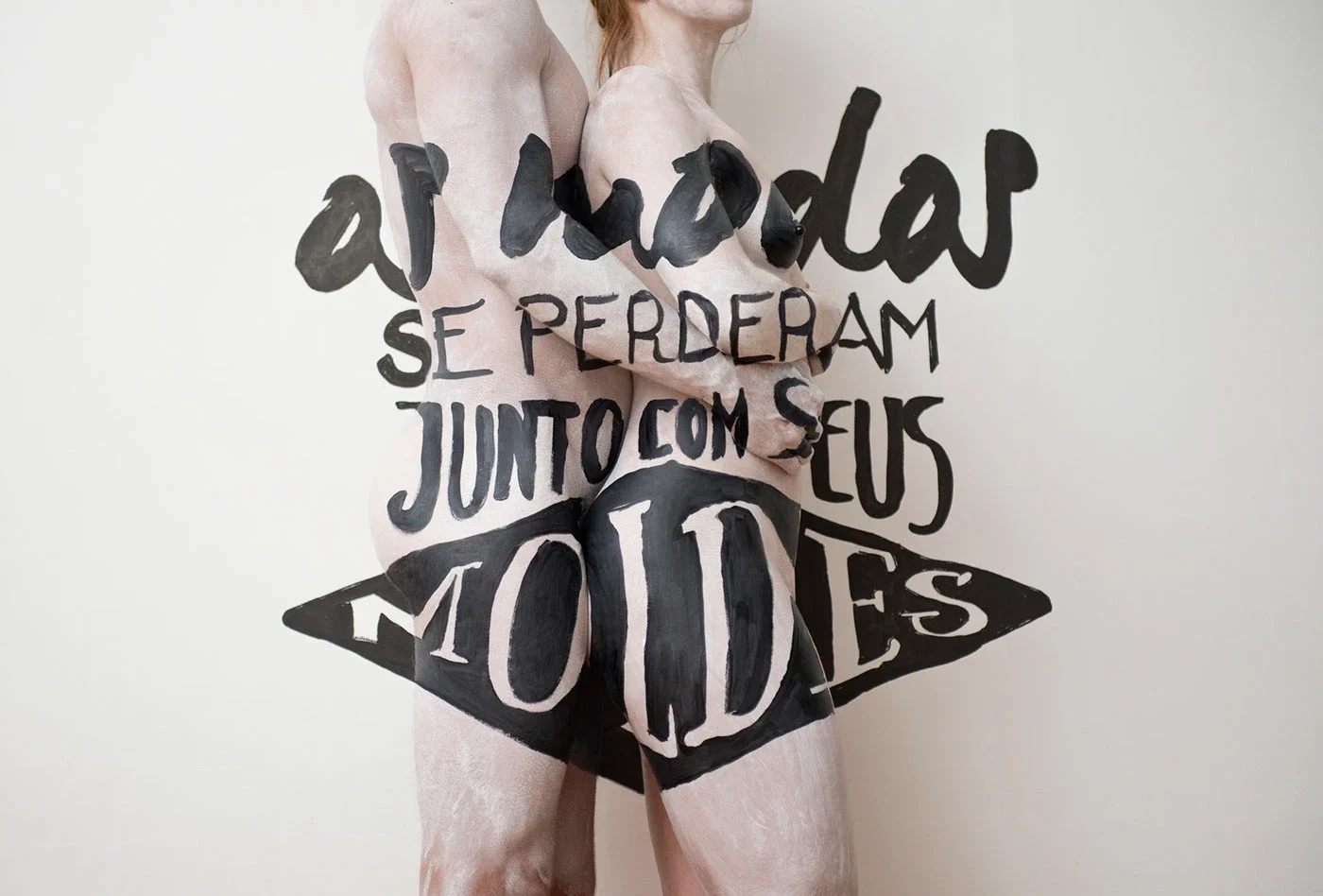 Duas pessoas nuas, de costas, com palavras em português pintadas no corpo e na parede, transmitindo uma mensagem de união e perda