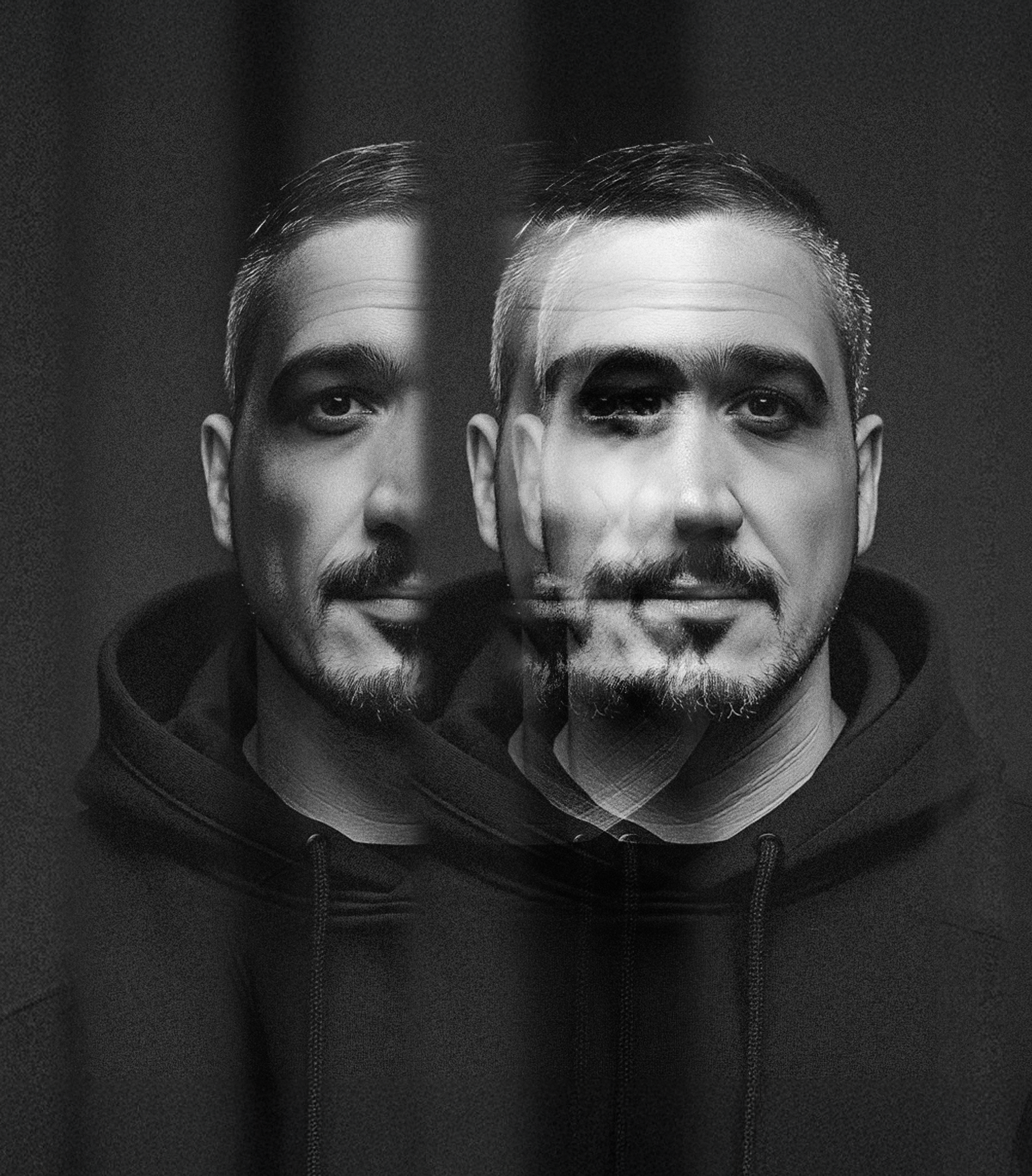 Foto em preto e branco de um homem com um espelho de efeito de ilusão, criando uma duplicidade no rosto, usando um hoodie preto.