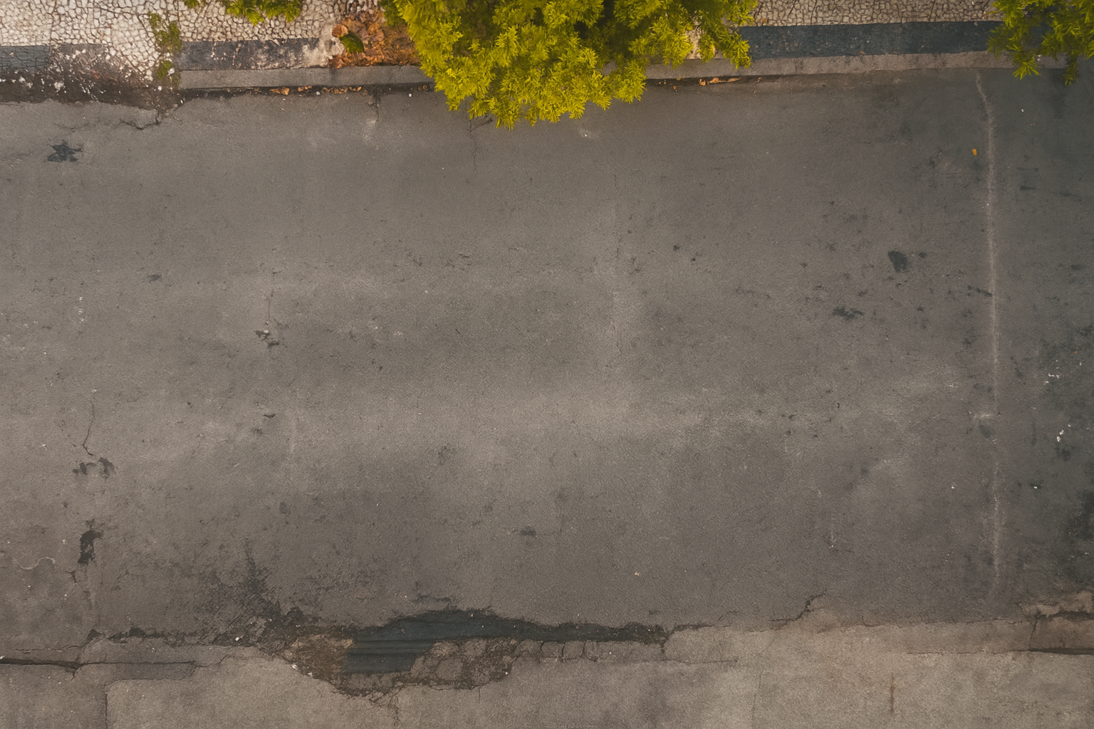 Imagem de uma calçada de rua de asfalto com alguns pontos de desgaste e uma borda de pedra ao longo de um muro com vegetação acima.