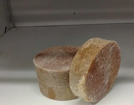 Two cylindrical brownish soap bars with a rough texture, placed on a white surface in a store shelf.