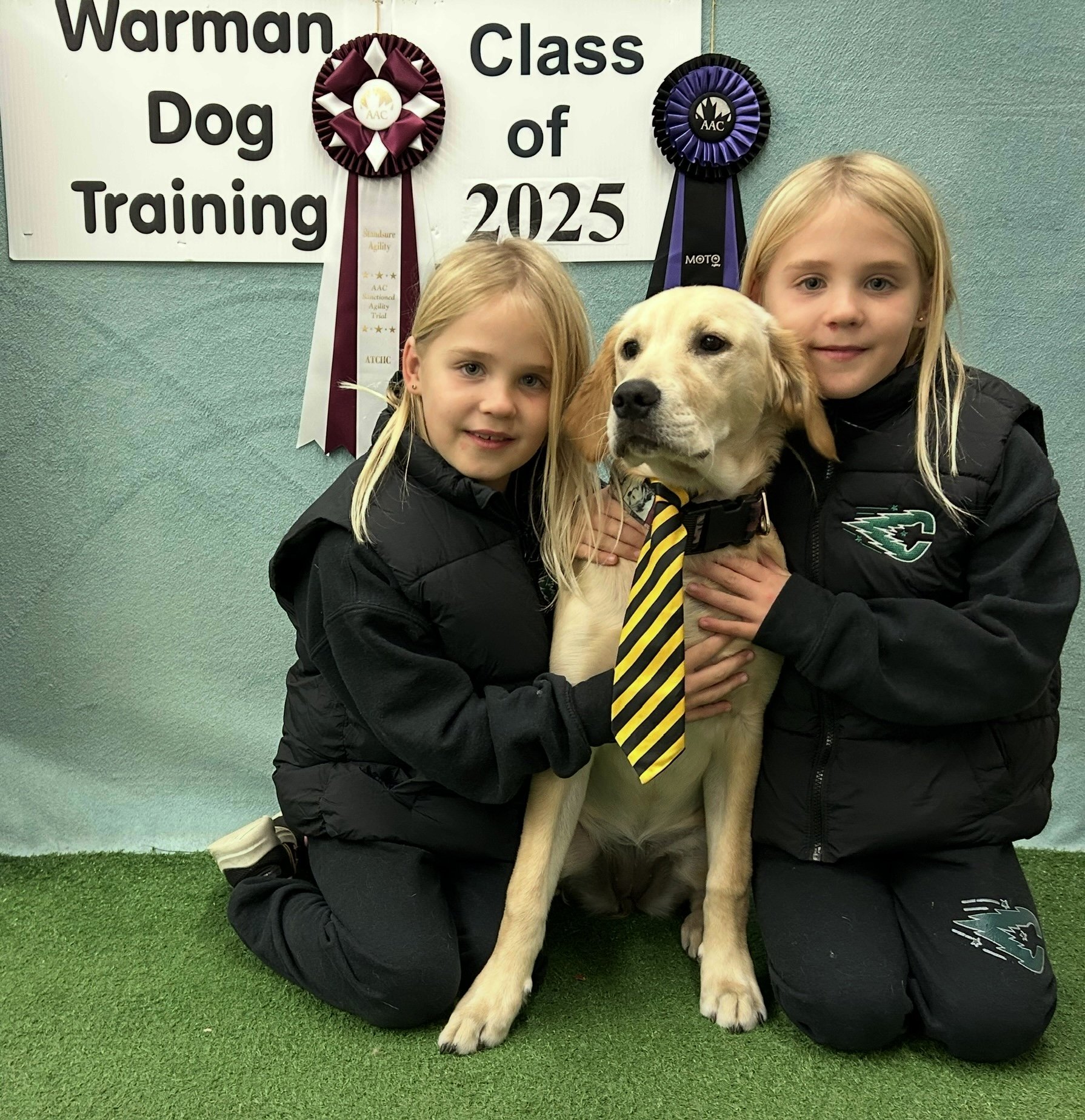 Two blond Girls hugging their lab puppy in front of Warman Dog Training graduation post.