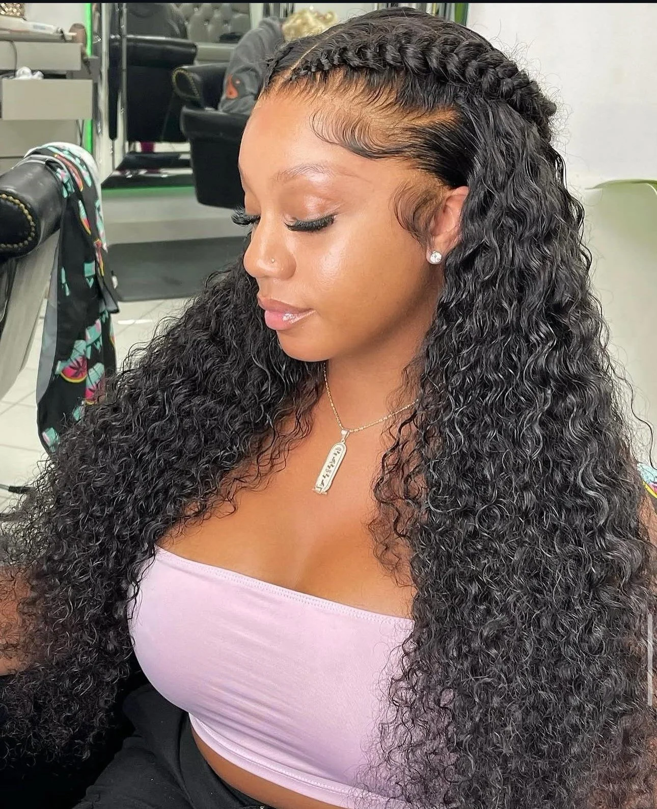 A woman with long, curly black hair and braids at the front, wearing a light pink tube top, sitting in a salon chair with a mirror and salon supplies in the background.