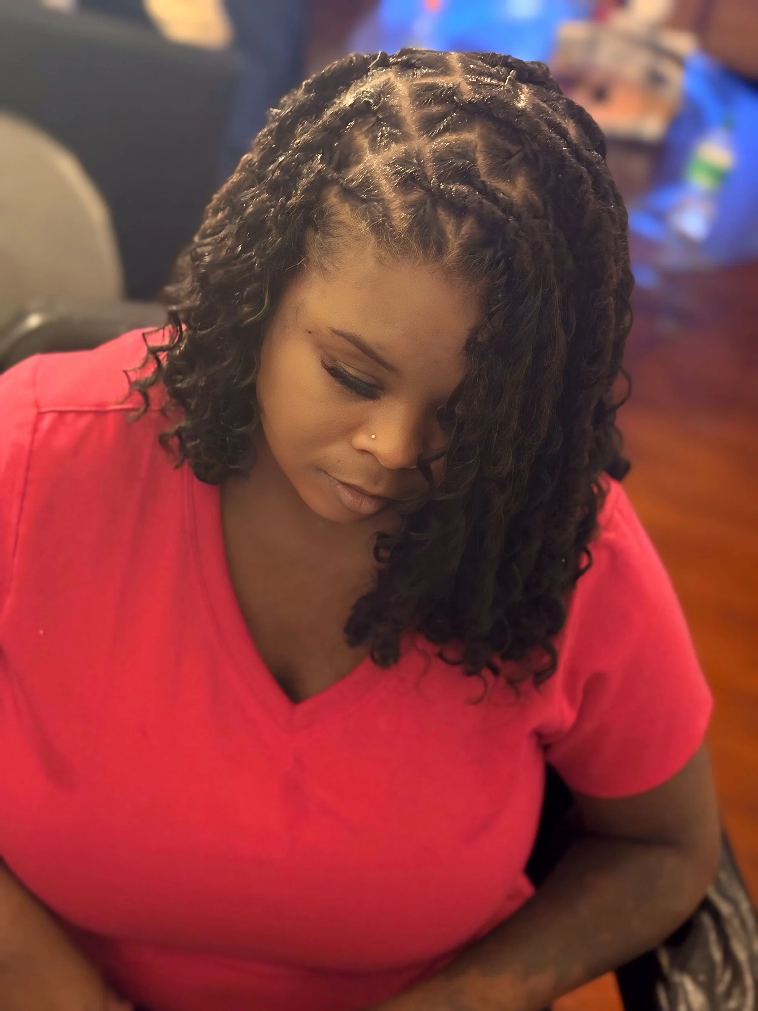 A woman with curly hair wearing a pink shirt, looking downward with a neutral expression.