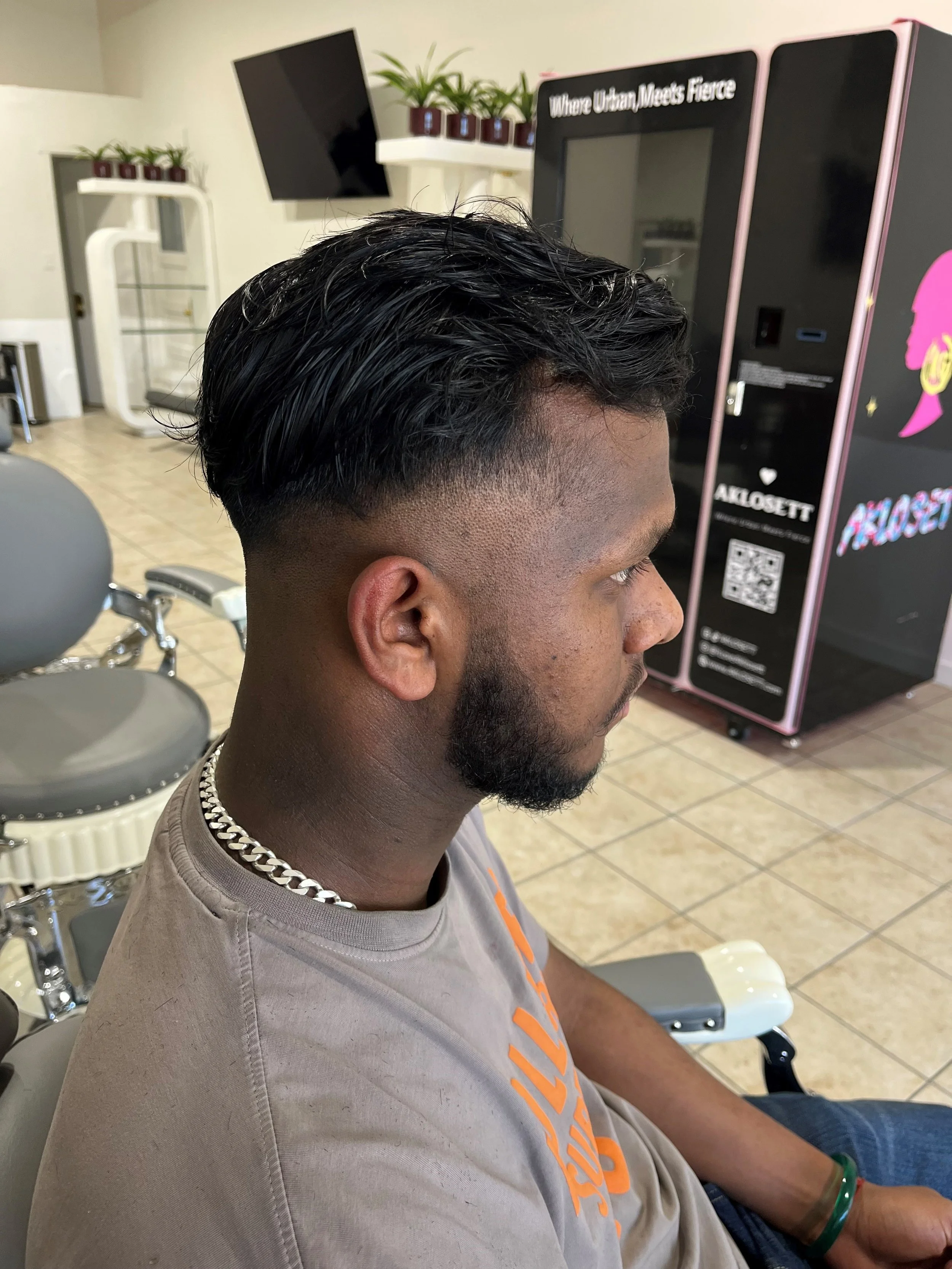 Side view of a man with black, wavy, slicked-back hair and a fade haircut, sitting in a barber shop with modern decor. He has a beard, is wearing a beige T-shirt with orange print, a silver chain necklace, and a green bracelet on his wrist.