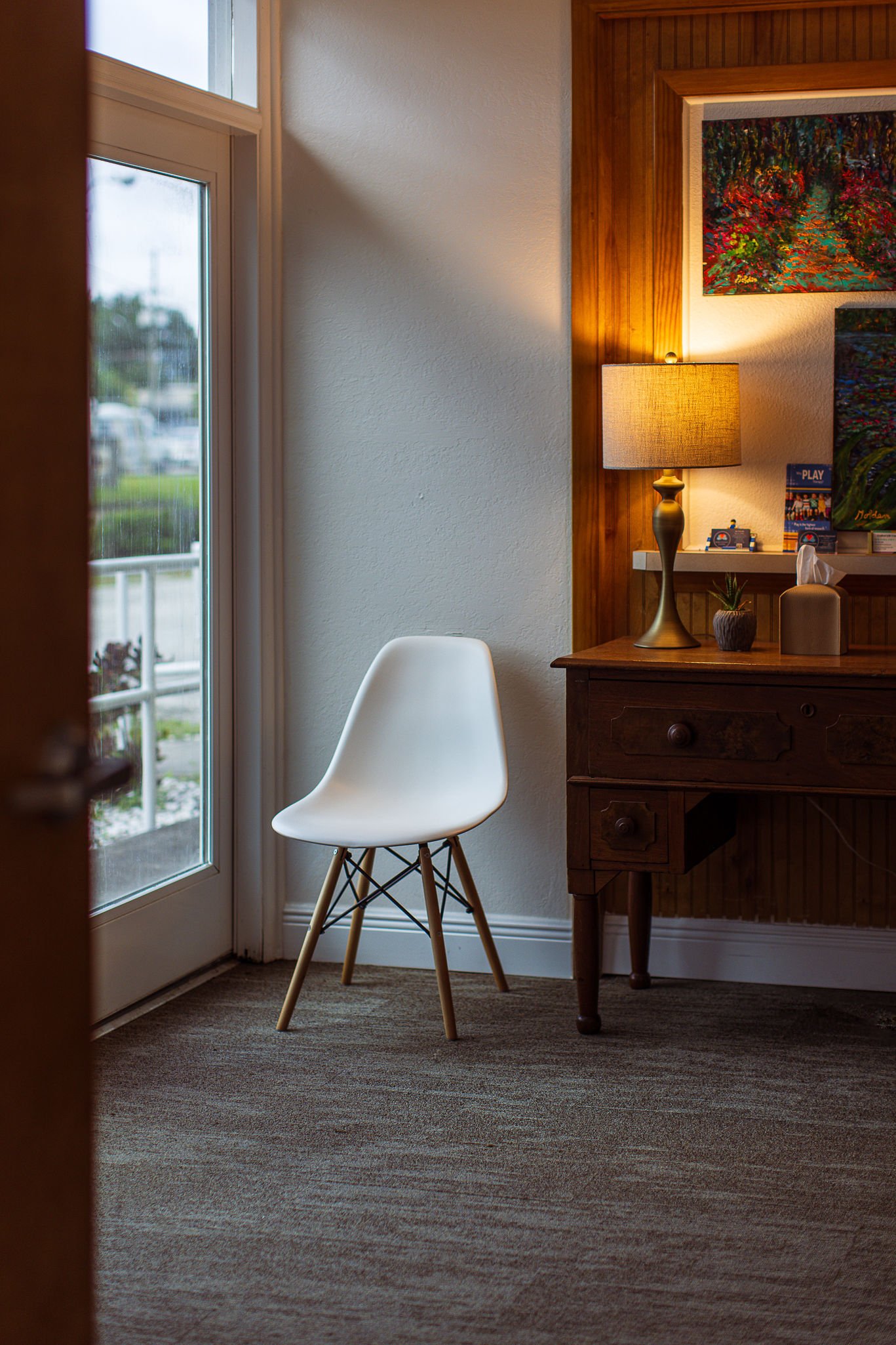 Interior scene with a white modern chair near a glass door, a wooden side table with a lamp, small plant, and tissue box, and artwork on the wall.