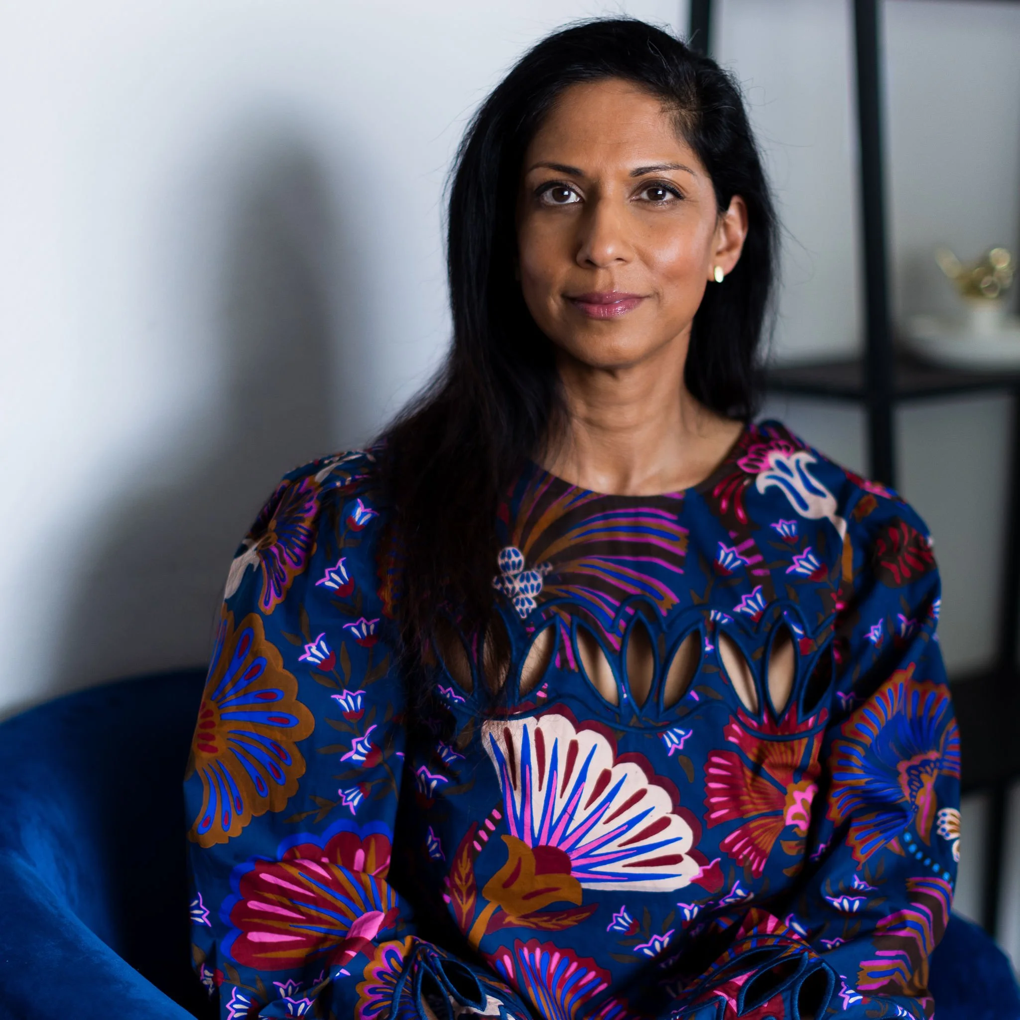 A woman with long black hair and medium skin tone posing indoors, wearing a colorful floral top with cutout details, sitting on a blue chair with a dark shelf in the background.