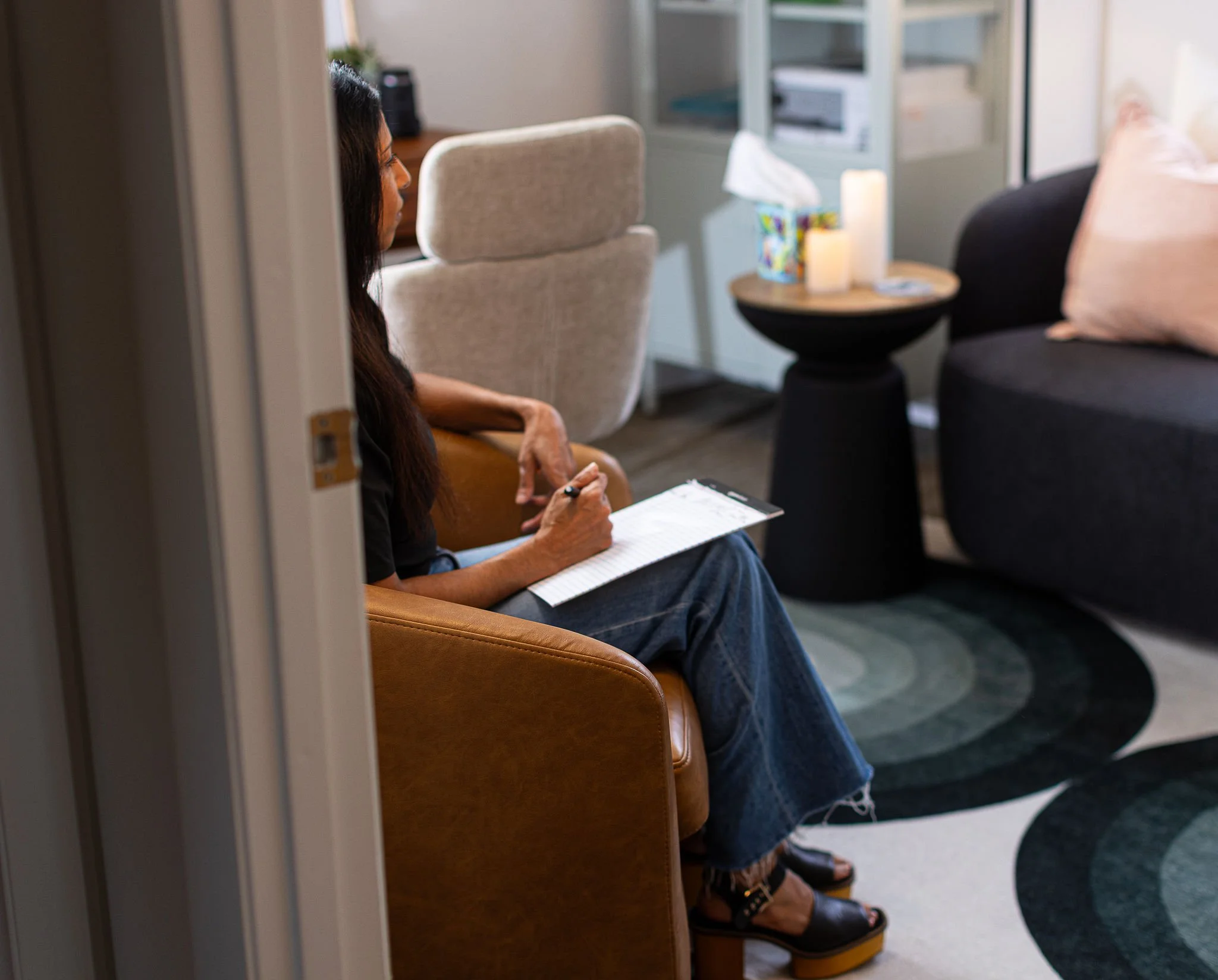 Woman sitting on a brown armchair, writing on a notepad in a cozy living room with a black sofa, a round black side table, and a white chair in the background.