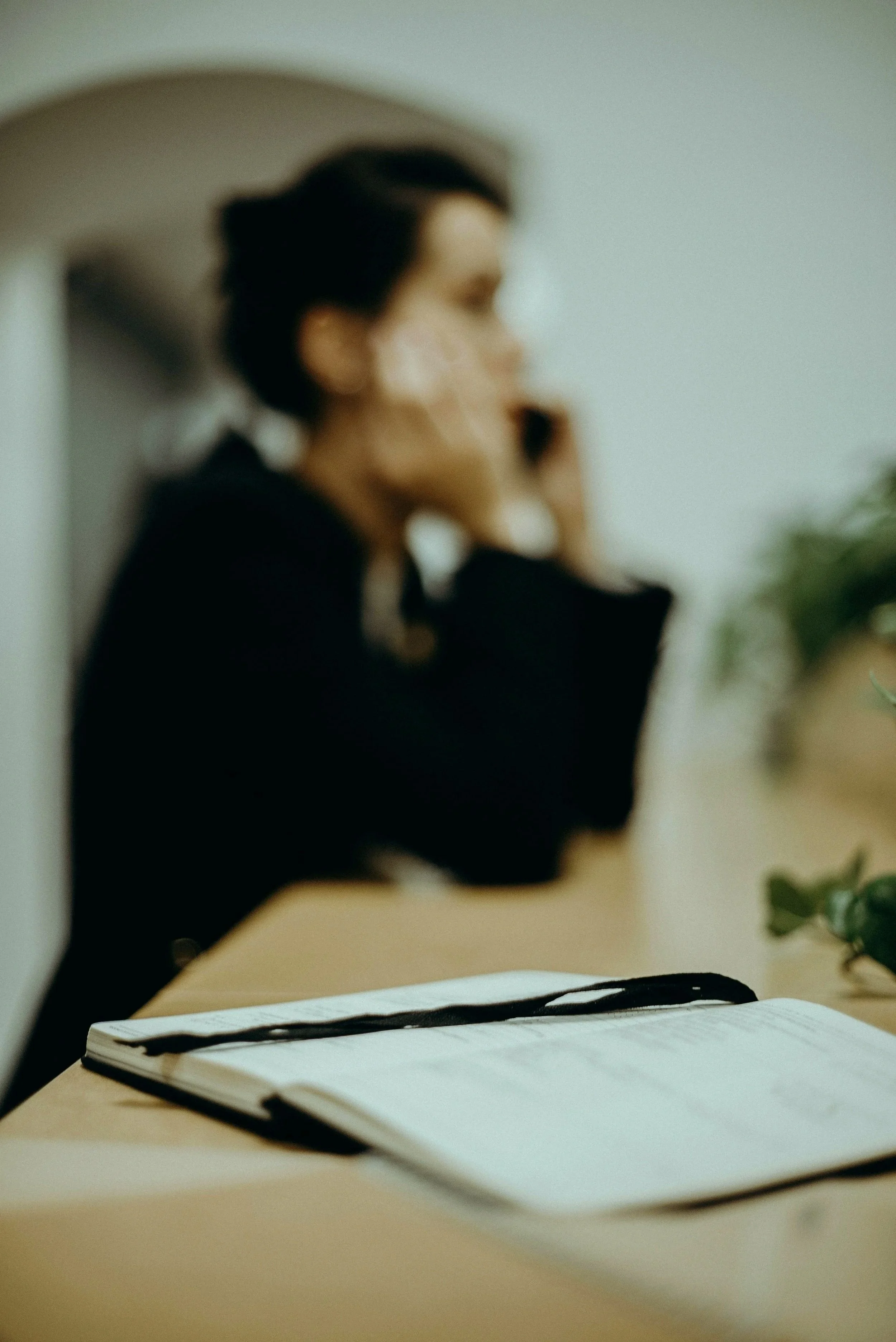 A blurred woman with dark hair sitting at a desk with an open notebook and a pen placed on top.