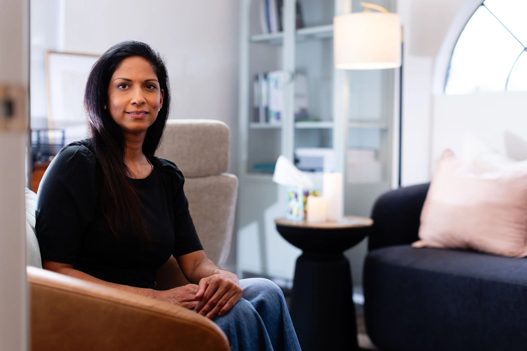 A woman sitting on a couch in a cozy, well-lit living room with modern decor, including a bookshelf, a side table with tissues, and a lampshade