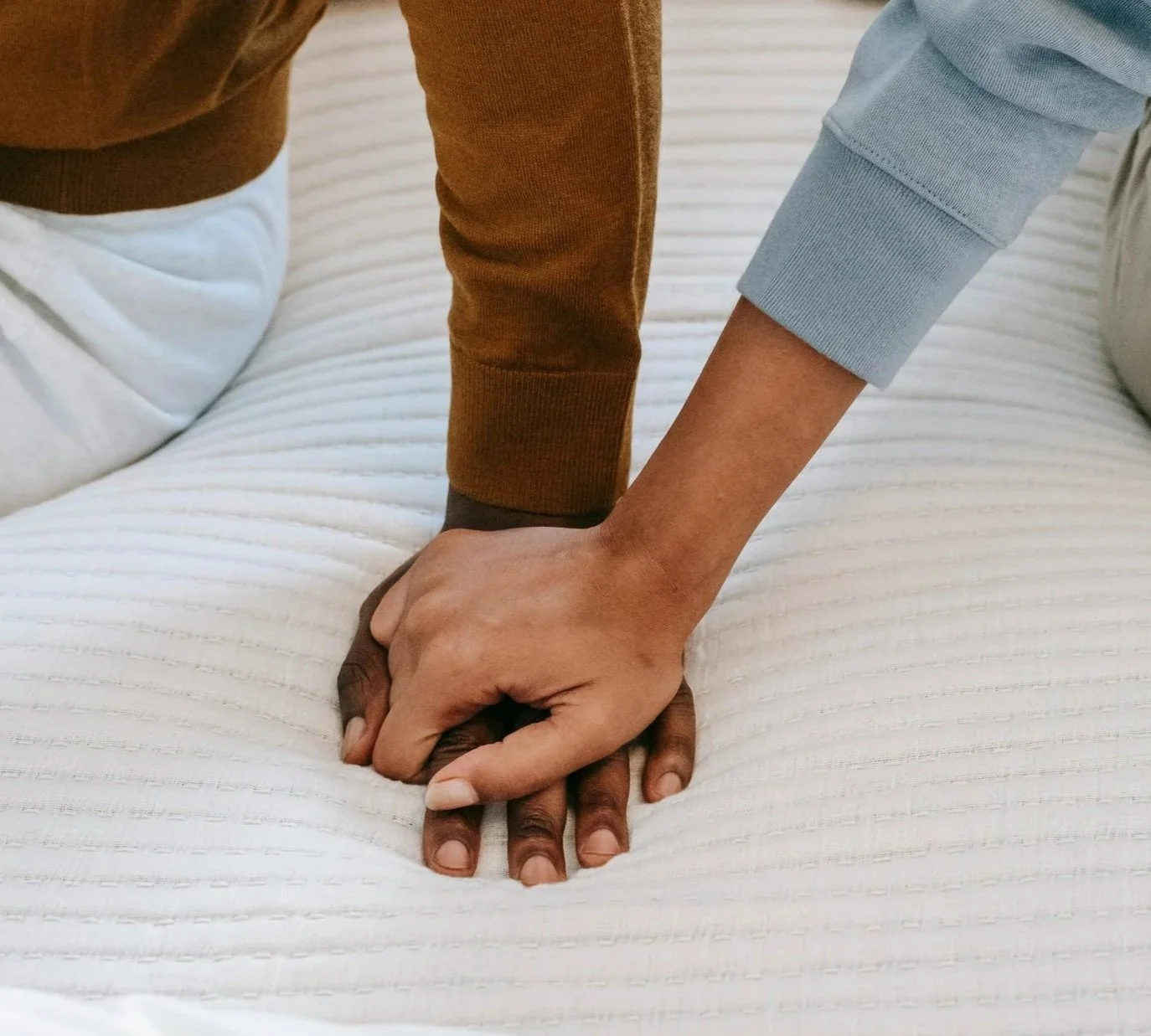 Two people holding hands on a white quilted bedspread, one person's hand grasping the other's.