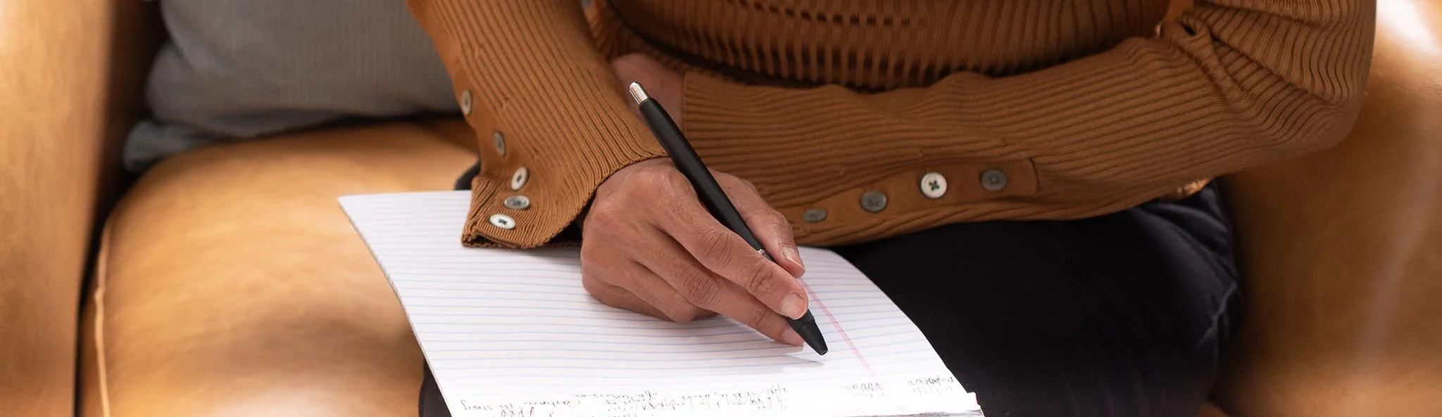 Close-up of a person wearing a brown jacket with buttons, writing on lined paper with a black pen while seated on a brown leather chair.