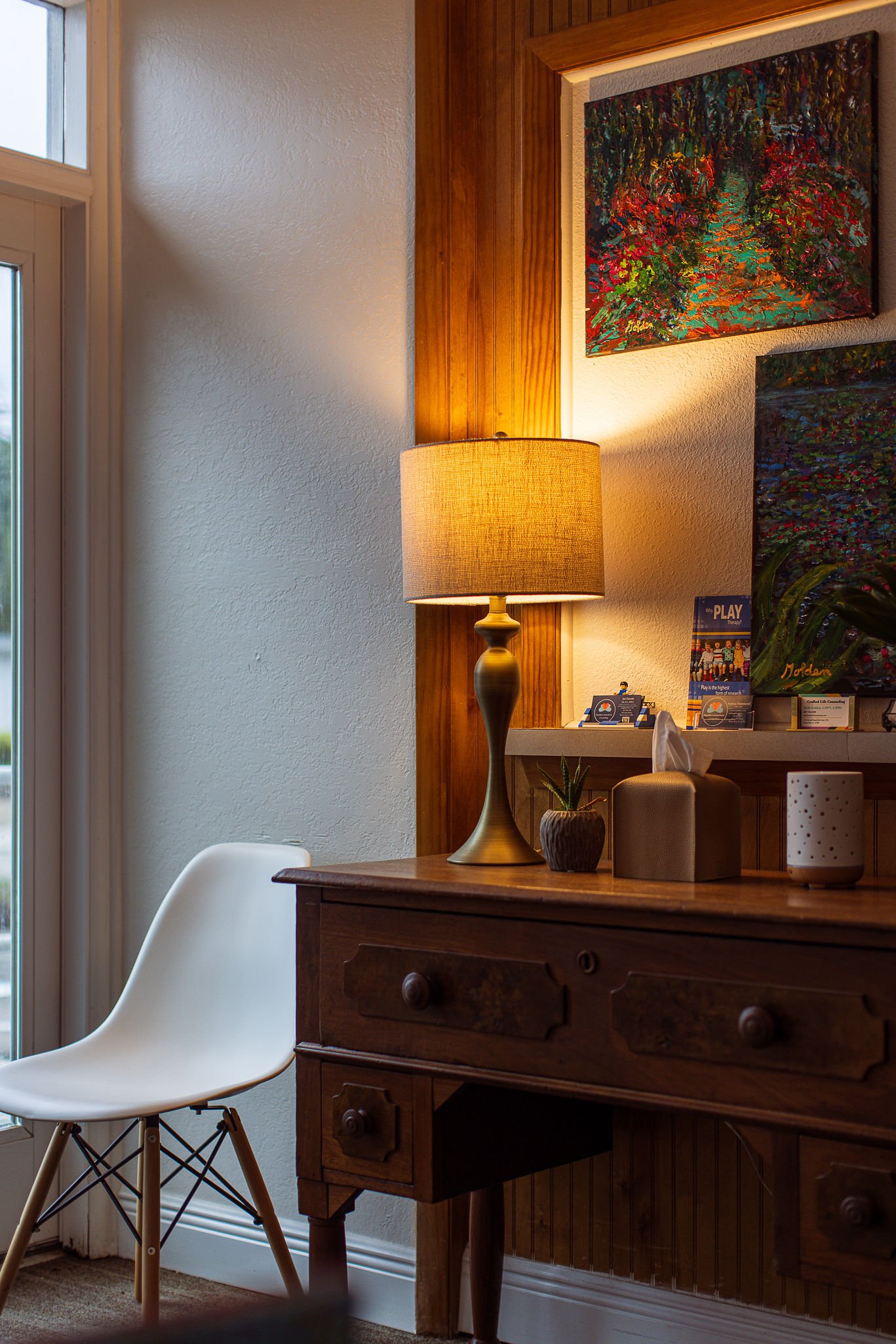 Interior view of a room corner with a window, a wooden desk, a white chair, a table lamp, and colorful paintings on the wall.