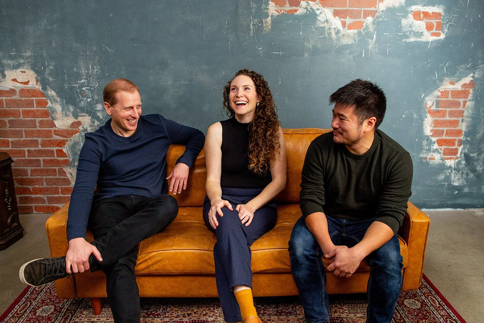 Three people sitting on a brown leather sofa against a distressed blue wall with exposed red brick. They are smiling and engaging in conversation.