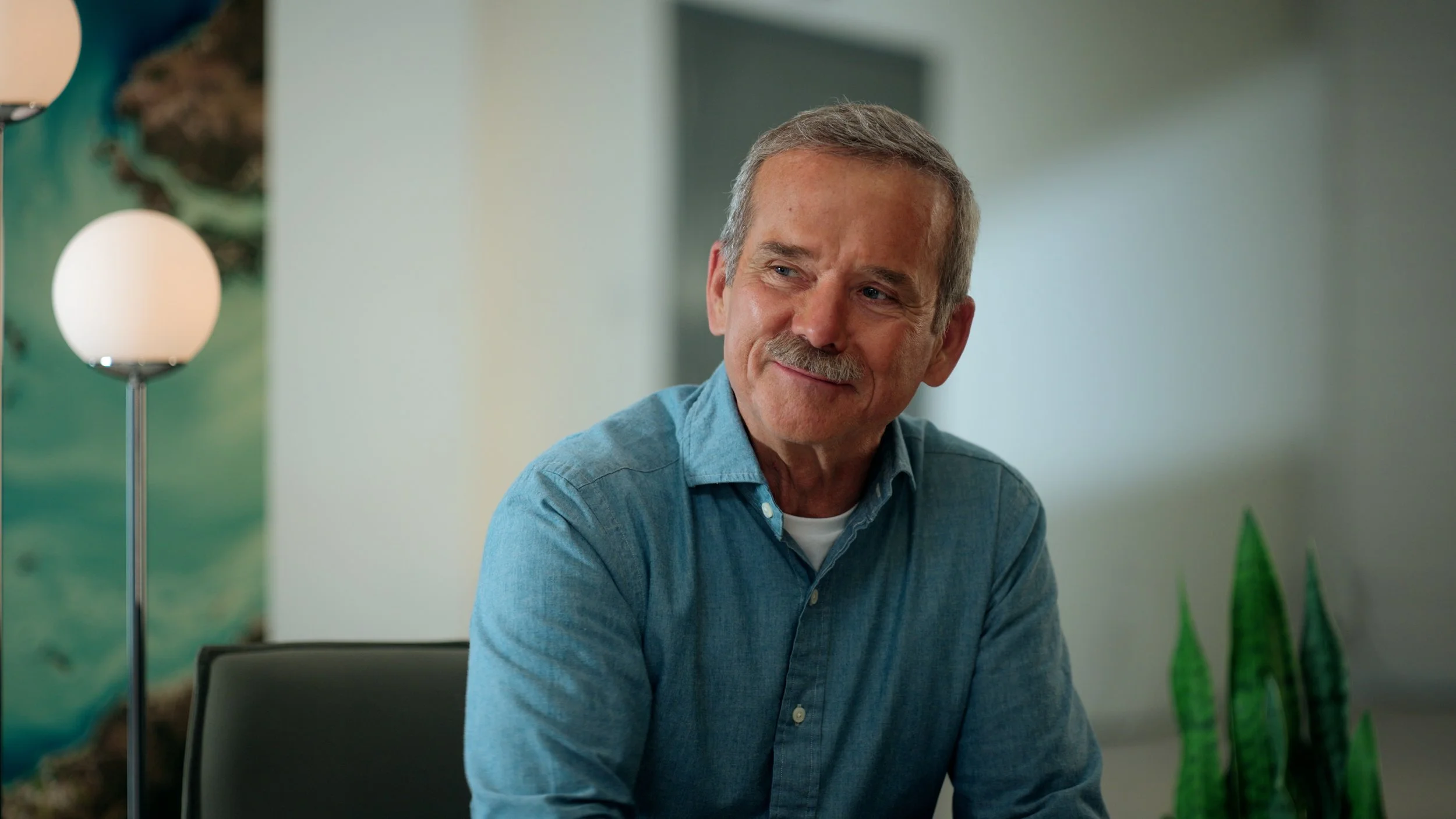 Chris Hadfield wearing a light blue button-up shirt, sitting at a table in a modern, well-lit room with a green plant on the right and a lamp with a round white shade on the left.