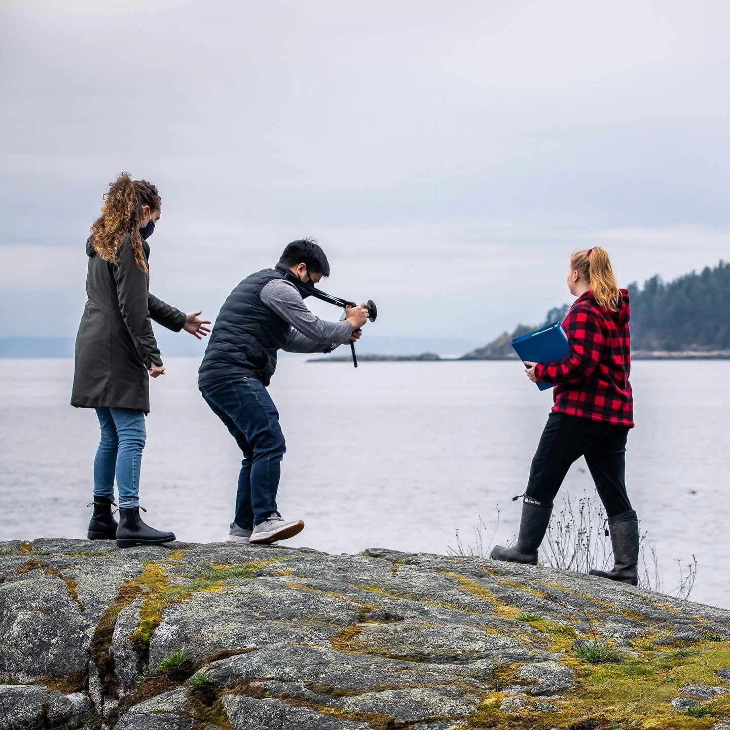 Three people standing on rocks near a body of water with a forested hill in the background. One person is filming with a camera stabilizer, while another is holding a blue folder, and the third is gesturing.