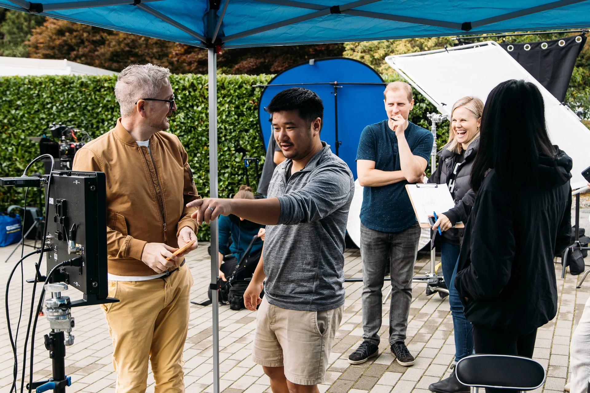 Filmmaking crew on outdoor set with lighting and film equipment, two women and three men talking, one man pointing at a monitor, greenery in background.