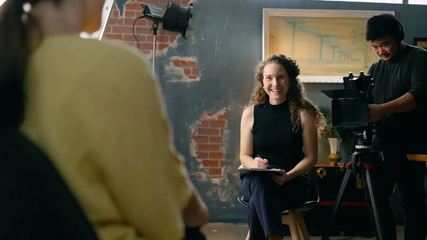 A woman smiling during an interview or recording session, sitting on a stool with a notepad and pen, with a camera and crew member filming her in a studio with a brick wall and artwork in the background.