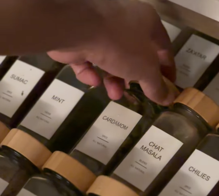 A hand reaching for a jar labeled 'Cardamom' among other spice jars labeled 'Sumac,' 'Mint,' 'Chaat Masala,' and 'Chilies.'