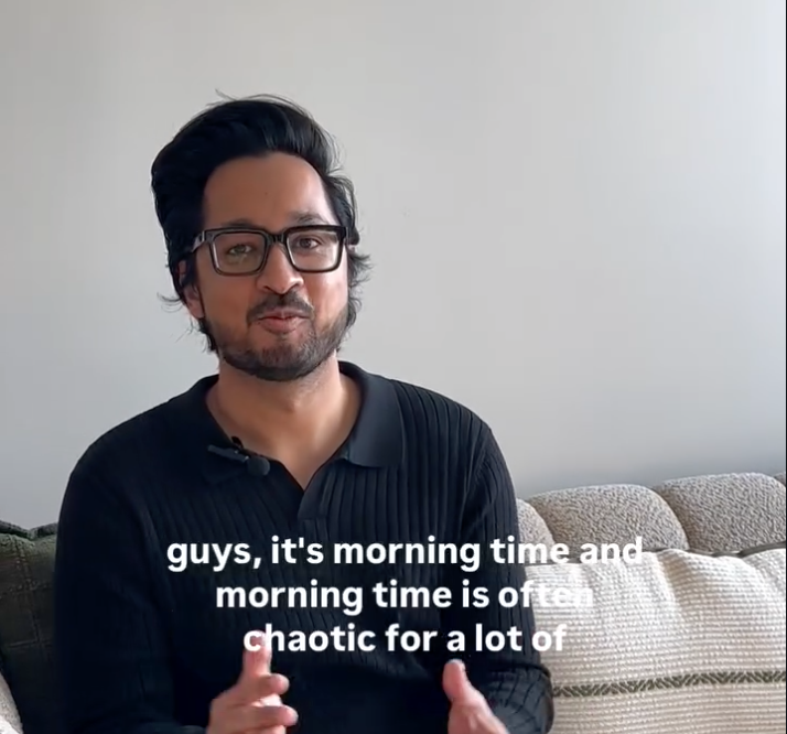 A man with dark hair and glasses sits on a beige couch in a room with a plain light-colored wall. He is wearing a dark sweater and speaking, with subtitles that read, 'guys, it's morning time and morning time is often chaotic for a lot of'.