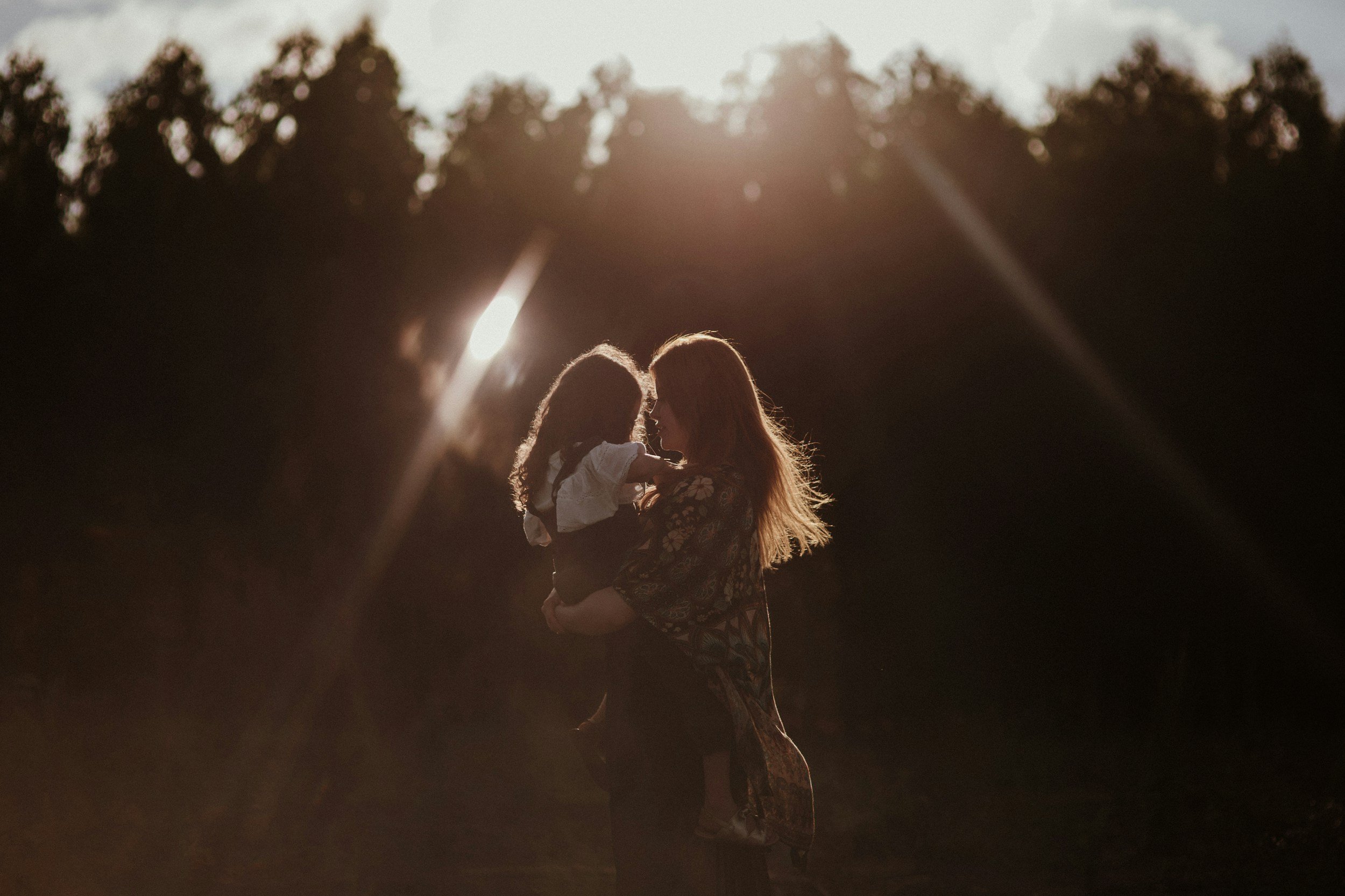 A woman holding a young girl in an outdoor setting during sunset, with trees in the background and sunlight creating lens flares.