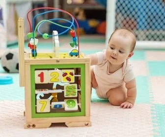 Baby playing with a wooden educational activity cube on a colorful play mat in a playroom with toys.