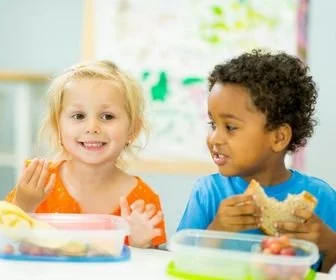 Two young children, a girl with blonde hair and a boy with curly black hair, sitting at a table with food containers, smiling and eating.