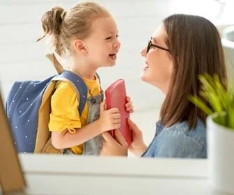 A young girl and a woman are smiling and facing each other indoors. The girl is holding a pink item, and the woman is wearing glasses. The background shows a white wall and a small plant.