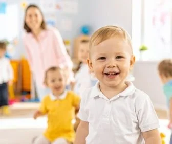 Smiling young boy in a white shirt in a bright, cheerful classroom with other children and a teacher in the background.
