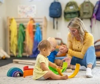 A woman and two young children playing with toys on the floor of a colorful indoor play area, with backpacks and jackets hanging on the wall in the background.