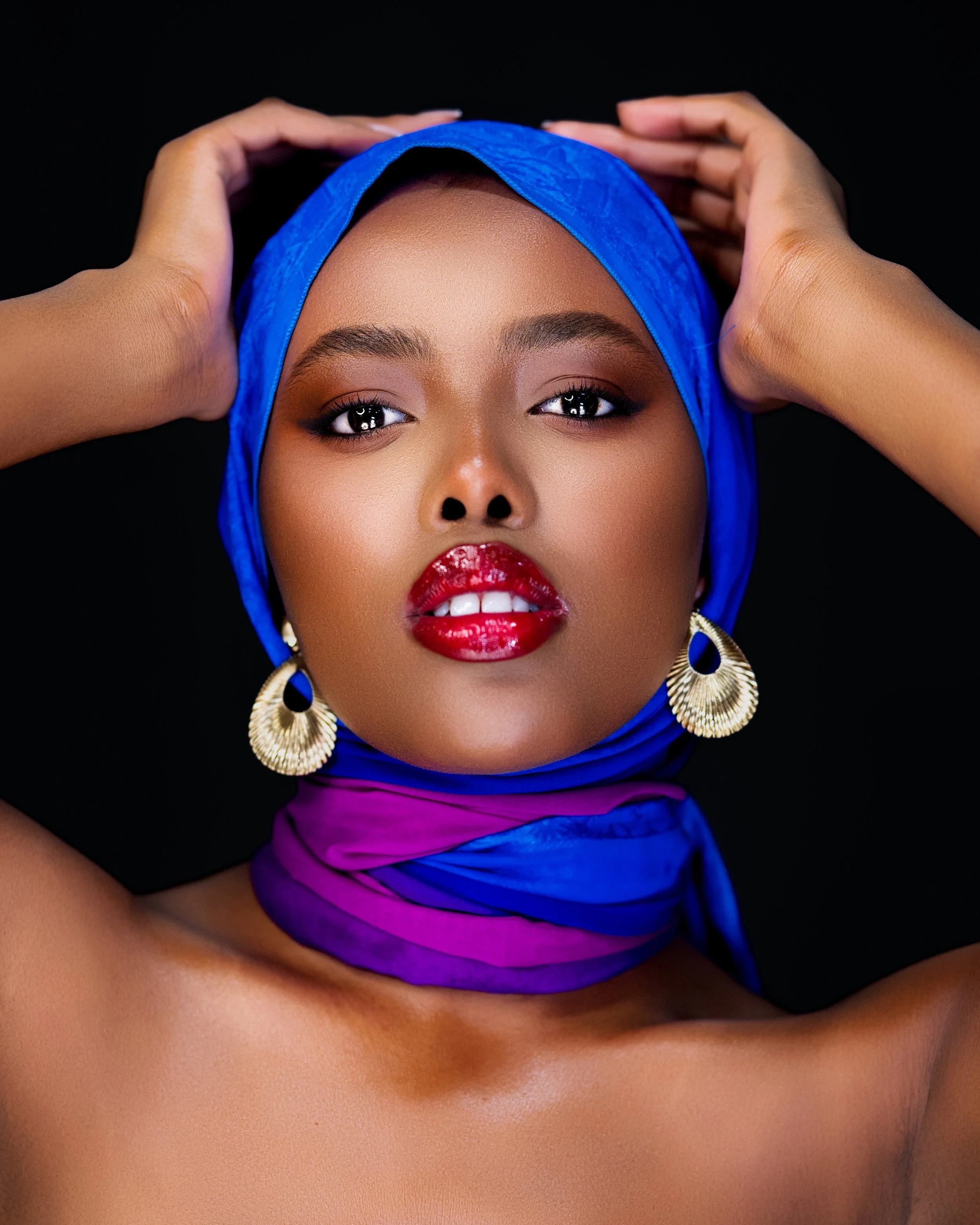 A woman wearing a blue headscarf and purple scarf, with bold red lipstick, gold earrings, holding her head with both hands against a black background.