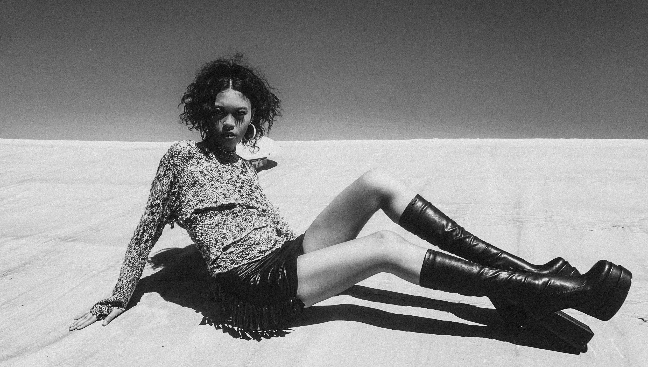 A woman sitting on the ground in a desert landscape, wearing a patterned top, leather shorts, and tall high-heeled boots. Black and white photo.