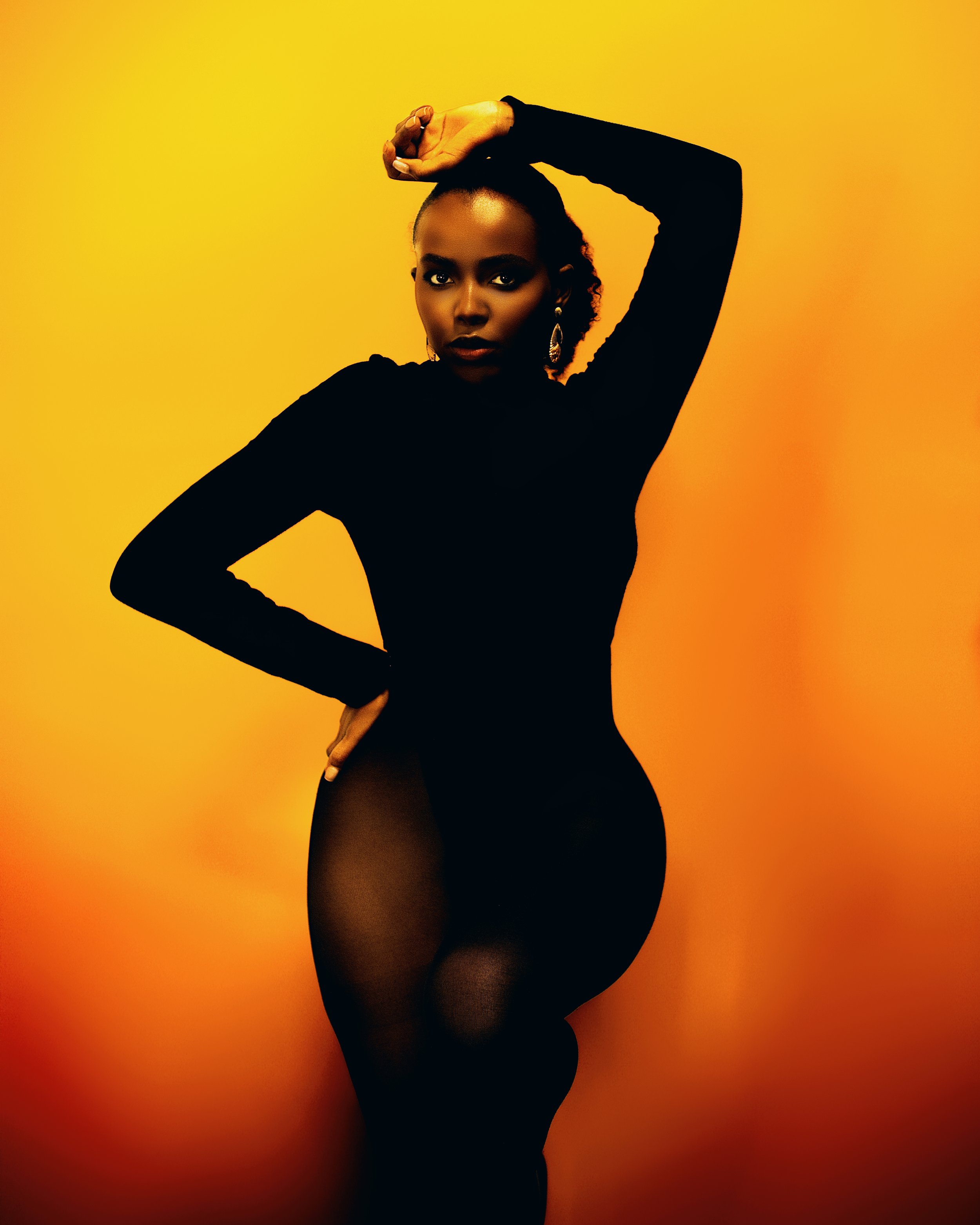 A woman in black clothing and sheer tights posing with one arm above her head and hand on her hip against a yellow-orange background.