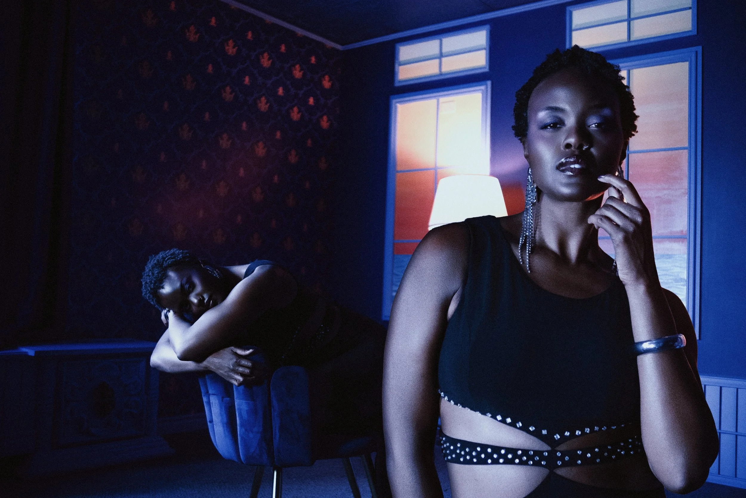 Two women in a dimly lit room with blue and purple tones; one is sitting on a chair and resting her head on her knees, the other is standing near a window, touching her chin with her finger, both dressed in black with jewelry.