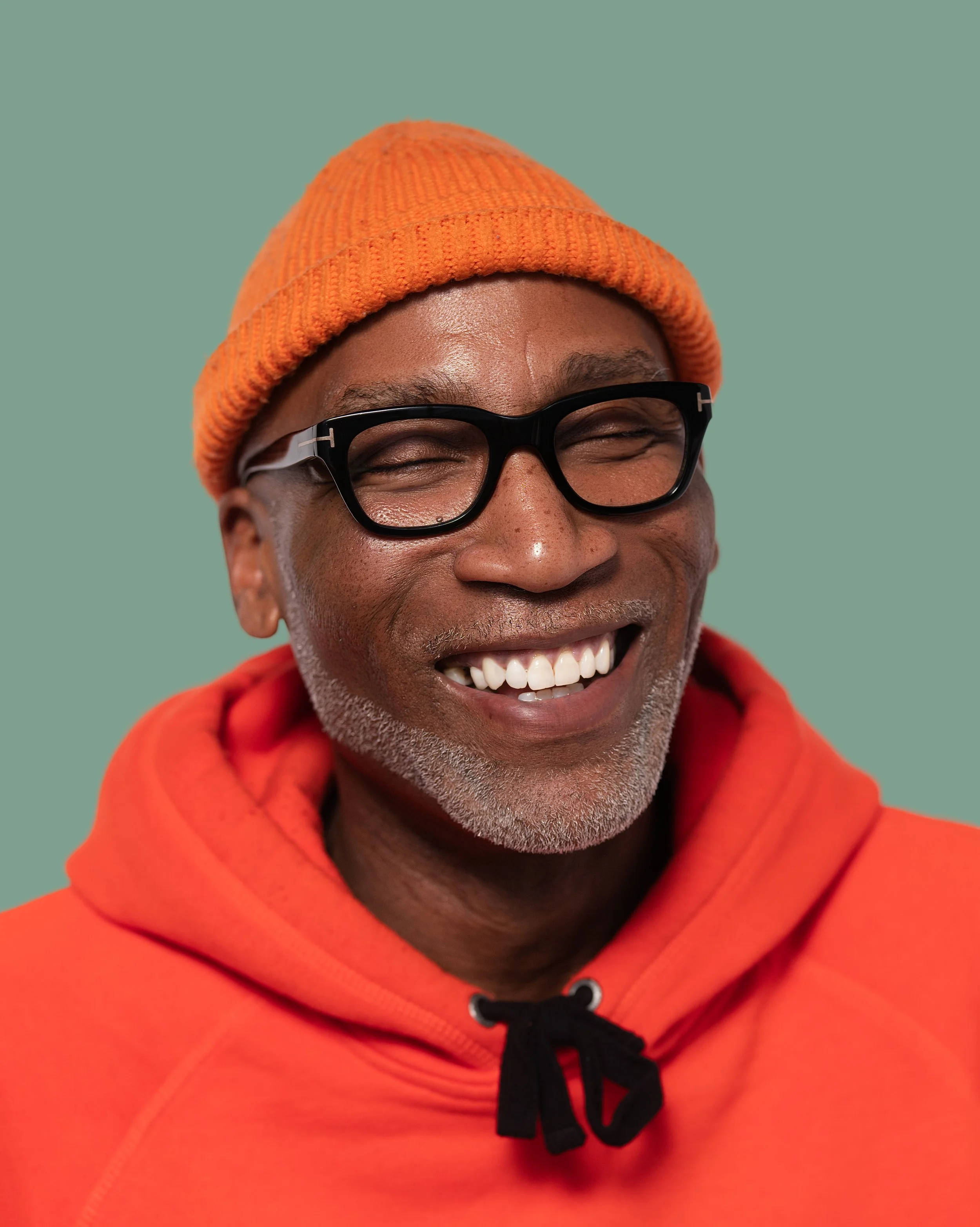 Smiling man wearing orange beanie, black glasses, orange hoodie, on a teal background.