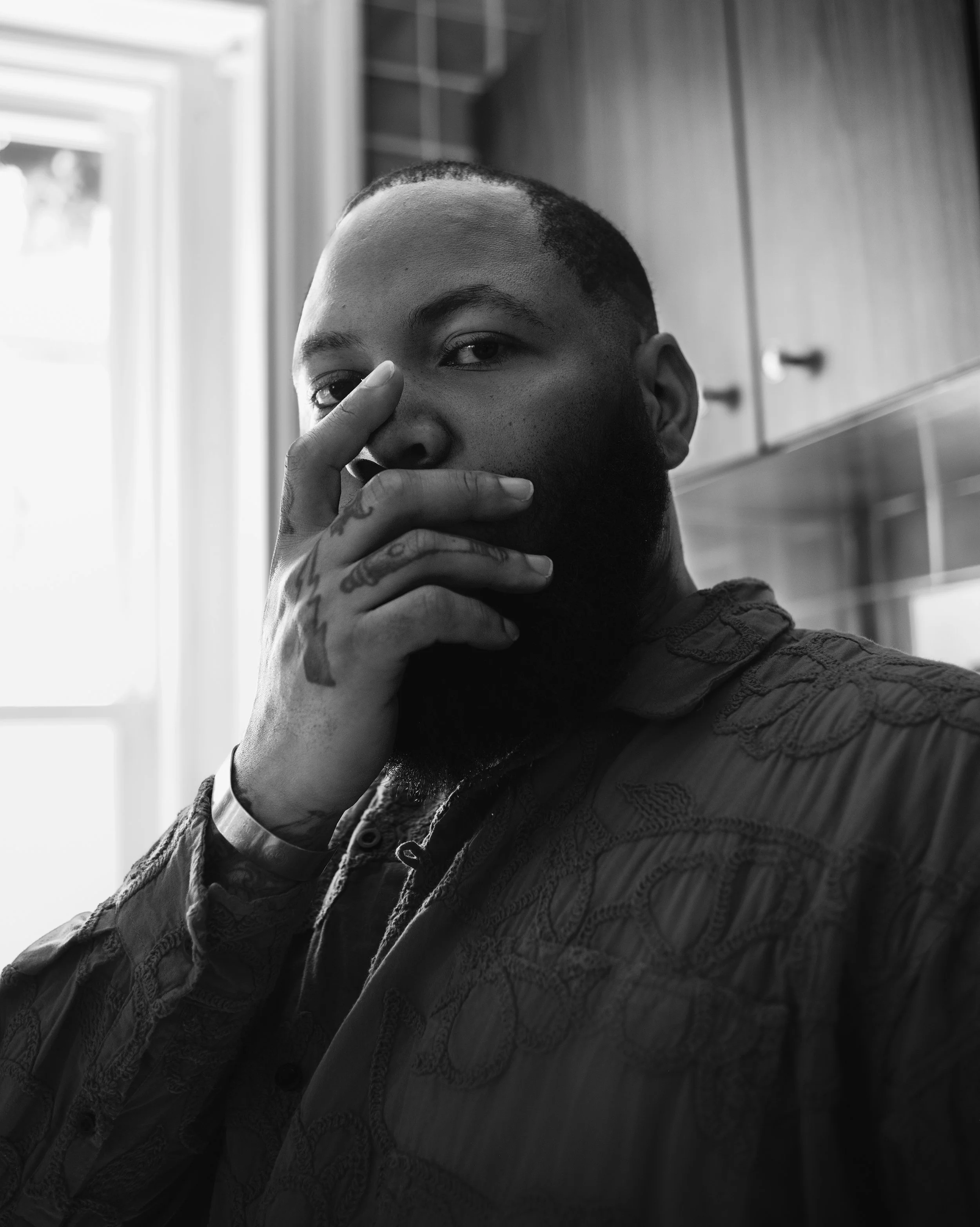 Black and white portrait of a man with a beard and tattoos, partially covering his face with his hand, standing in a room with natural light from a window.