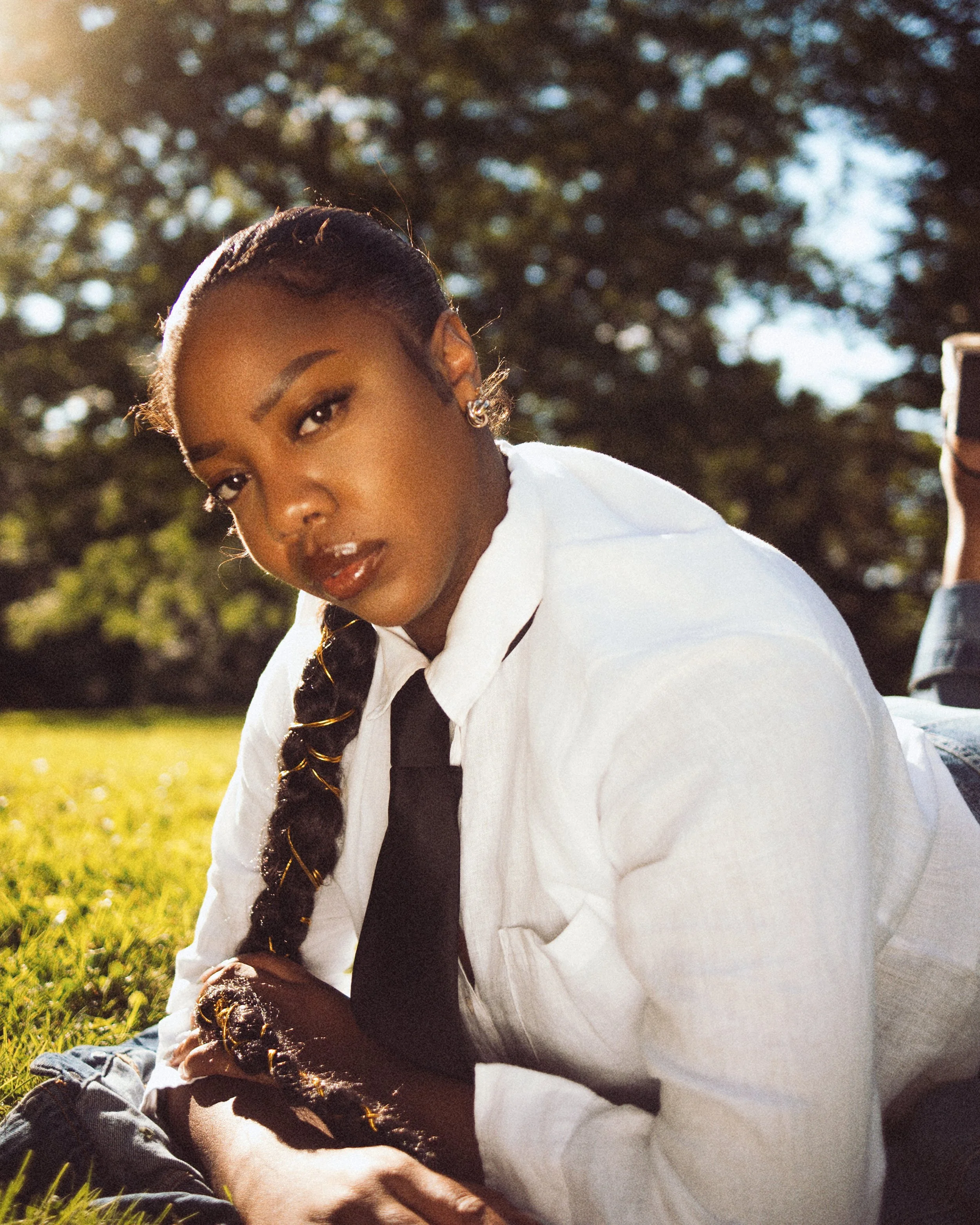 A woman with dark hair styled in a braid, lying on grass outdoors in sunlight, wearing a white shirt and black tie.