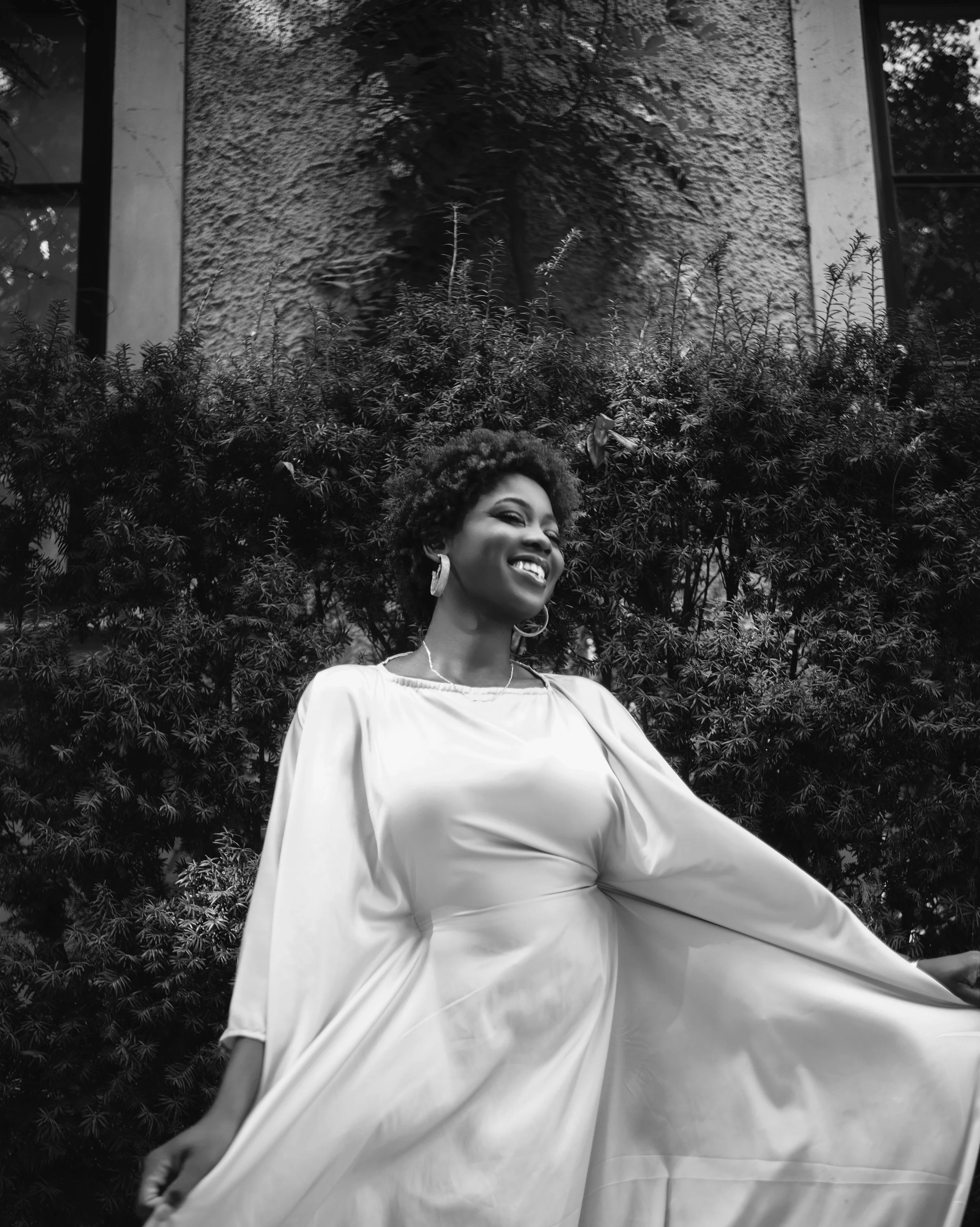 A woman with curly hair smiling in a flowing dress, standing in front of a bushy background, in black and white.