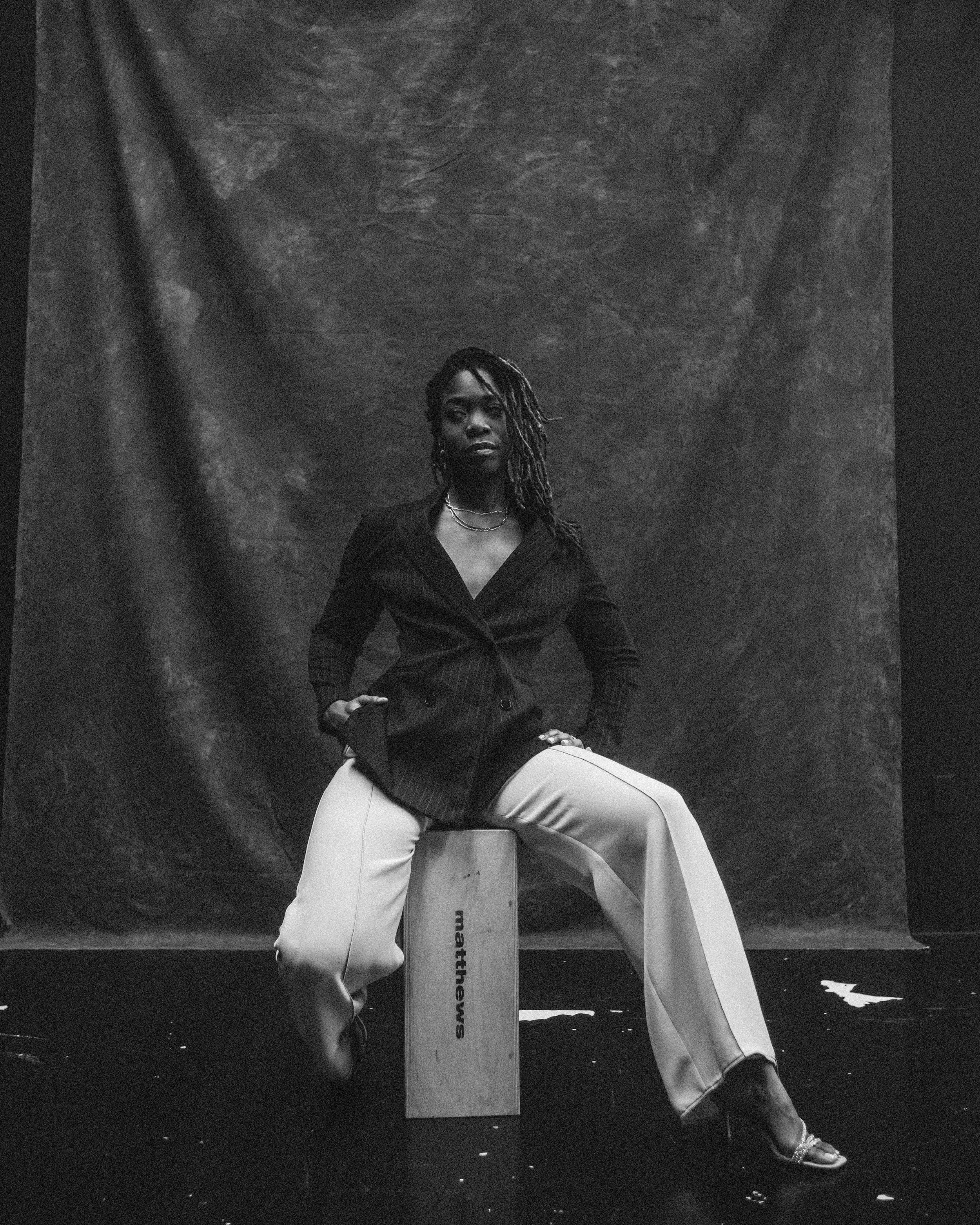 A woman with dreadlocks sitting on a wooden block in a photography studio. She is wearing a dark pinstripe blazer and light-colored wide-leg trousers, with jewelry and high-heeled sandals. The background is a dark textured backdrop.