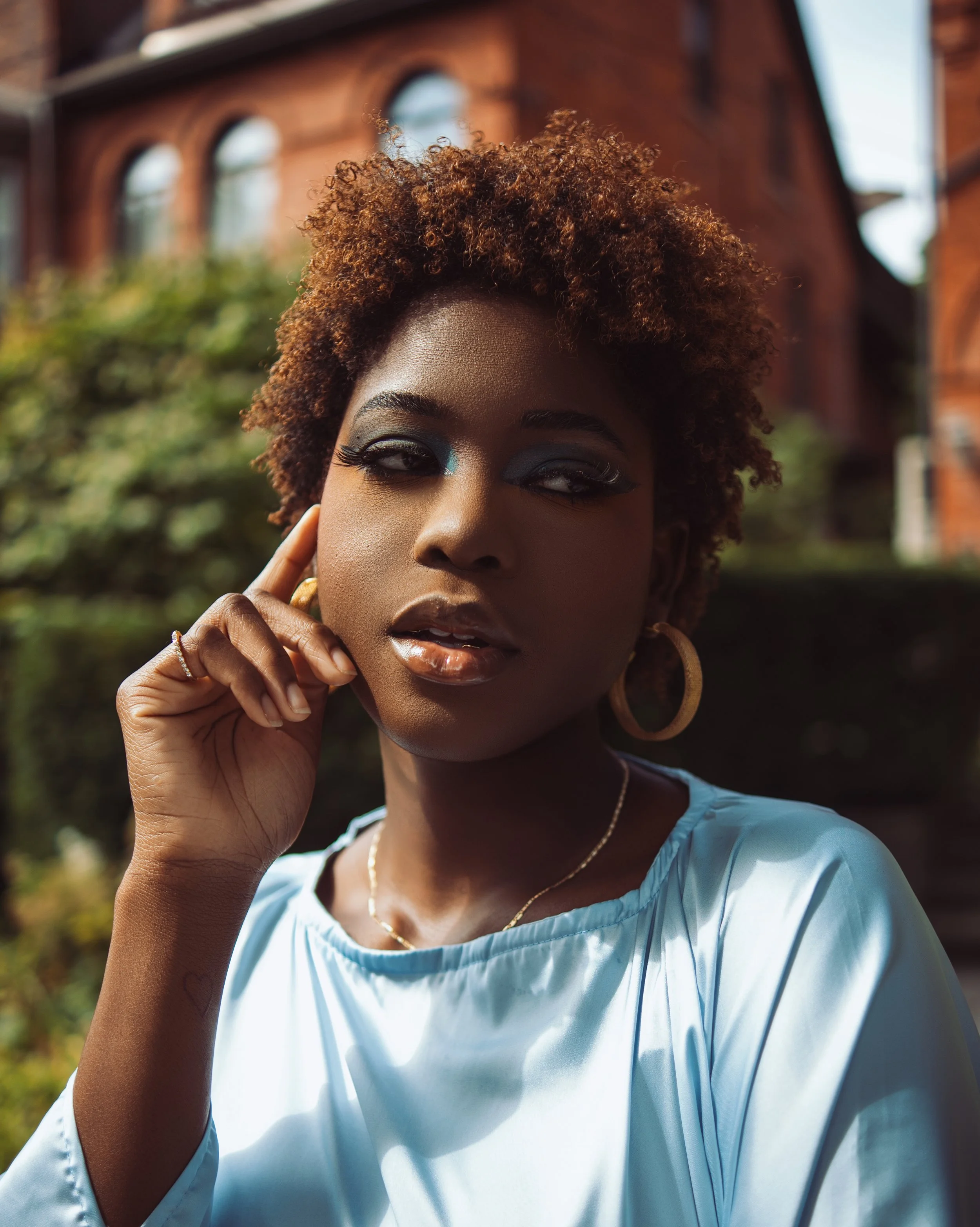 Portrait of a woman with dark skin and curly hair, wearing gold hoop earrings, a light blue satin top, and gold jewelry, standing outdoors in front of a red brick building and green bushes.