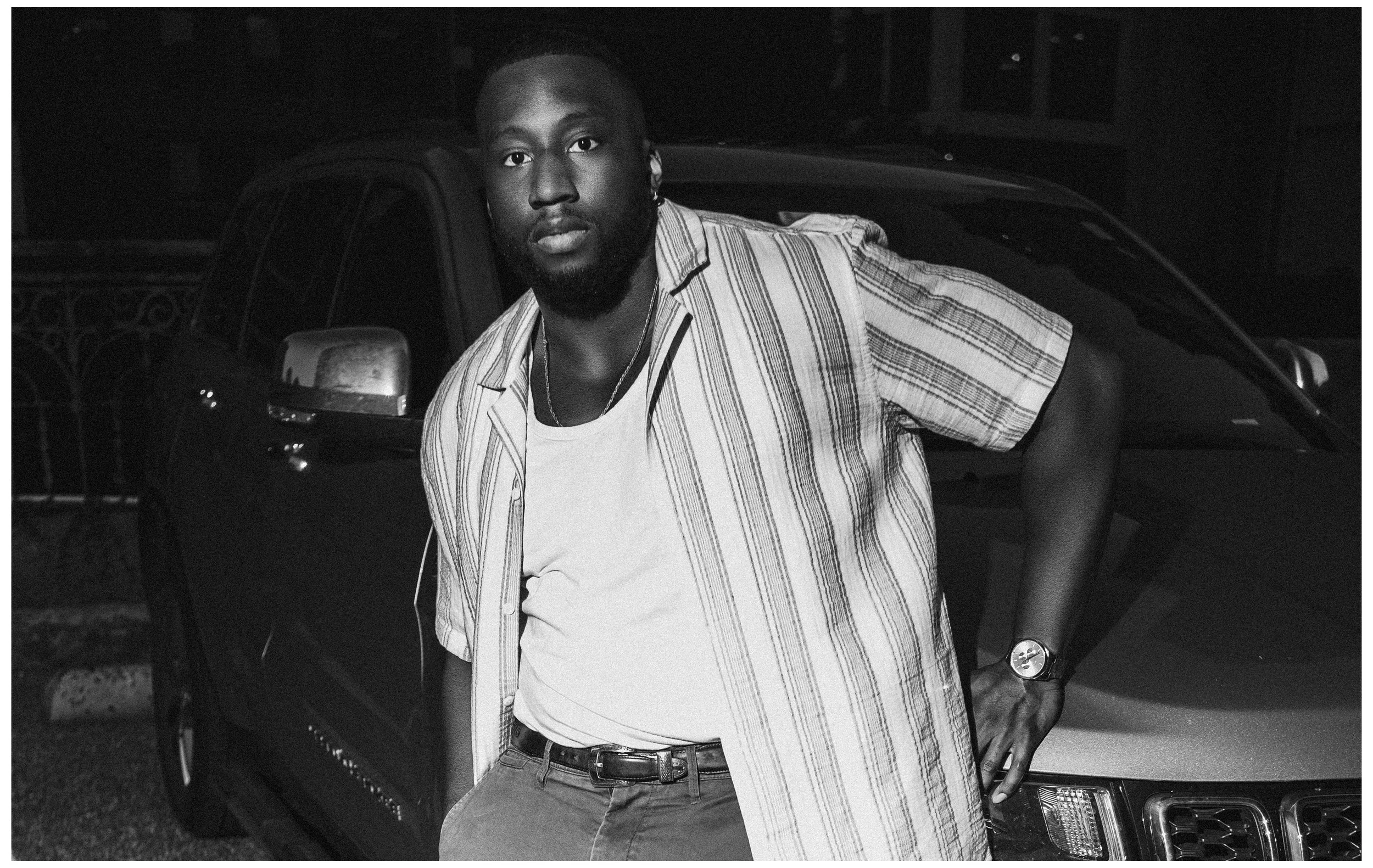 A man in a striped short-sleeve shirt and t-shirt leaning against a car at night.