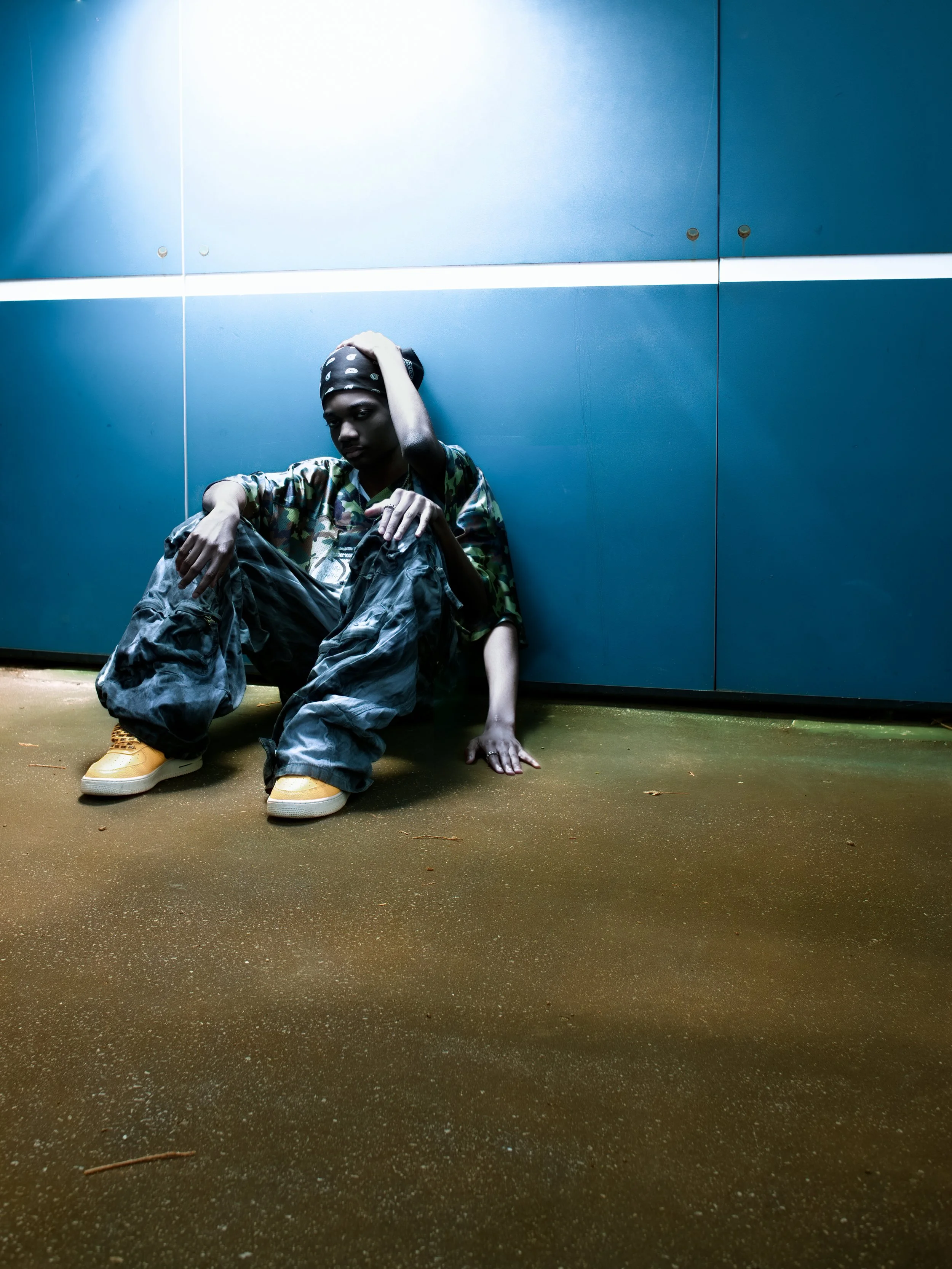 Person sitting on the floor against a blue wall, wearing camouflage shirt, black cargo pants, yellow sneakers, and a black bandana, with a contemplative expression.