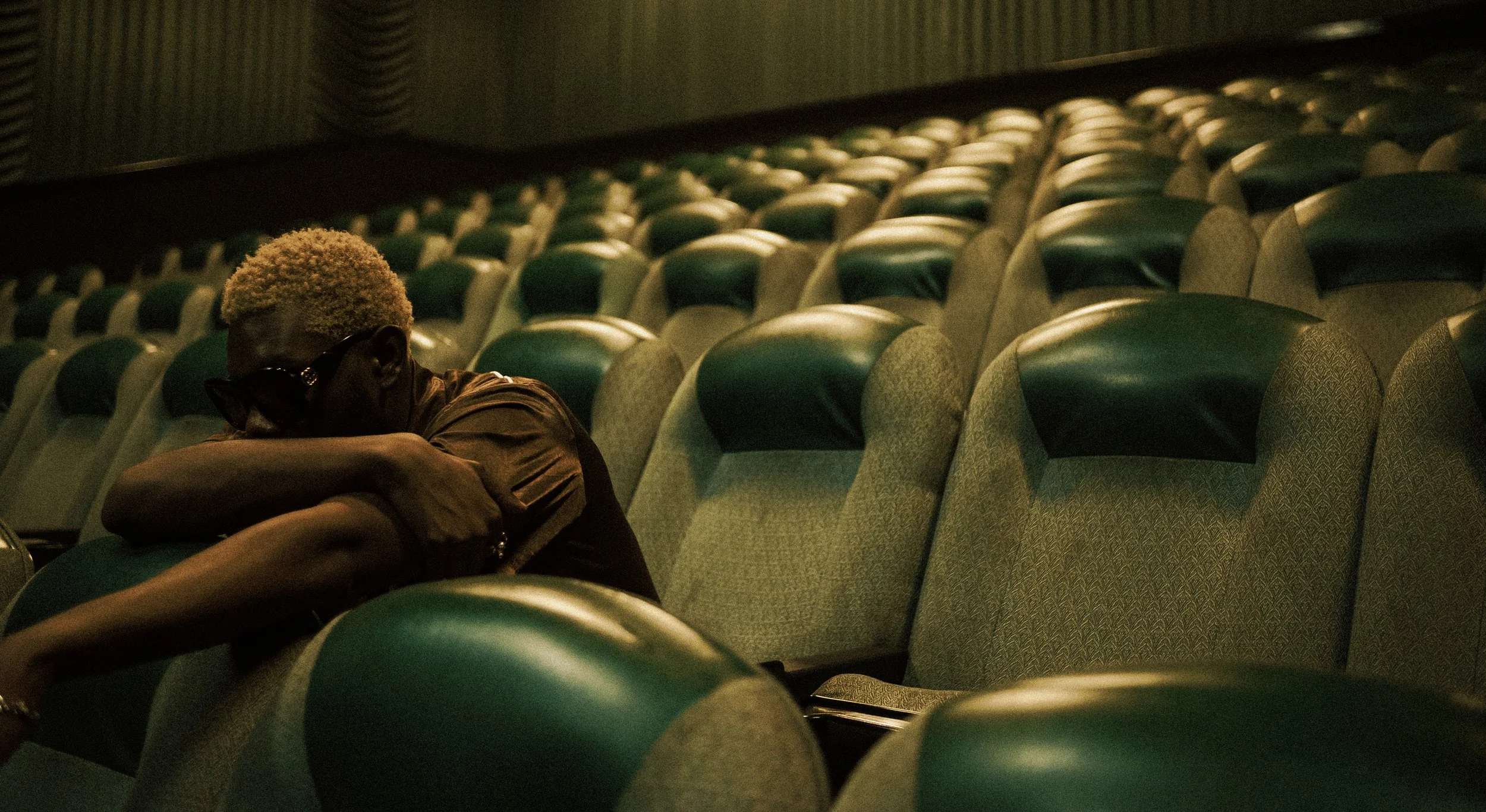 A person with light blonde hair and dark sunglasses sitting alone in an empty movie theater, resting on the armrest with cushions, the theater seats are green with black headrests, and the lighting is dim.