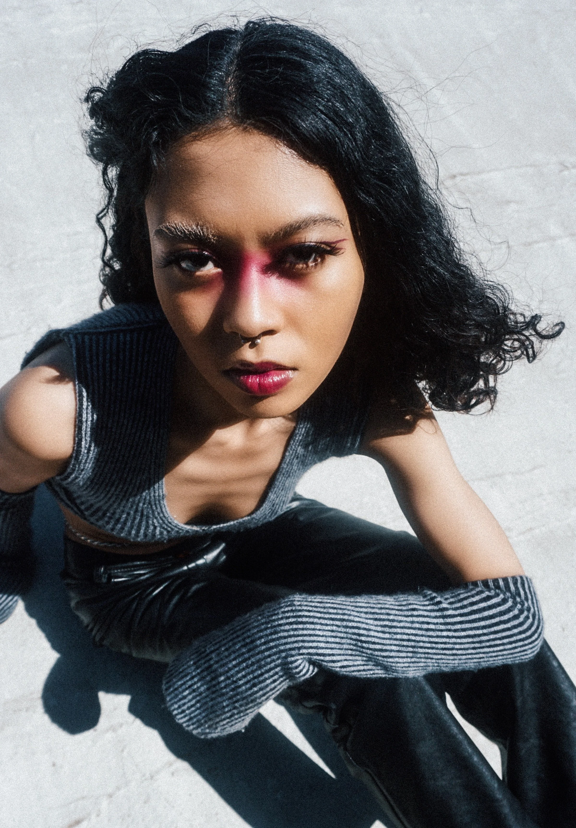 A young woman with curly black hair and bold makeup, wearing a sleeveless top and leather pants, crouching on a white surface, looking directly at the camera.