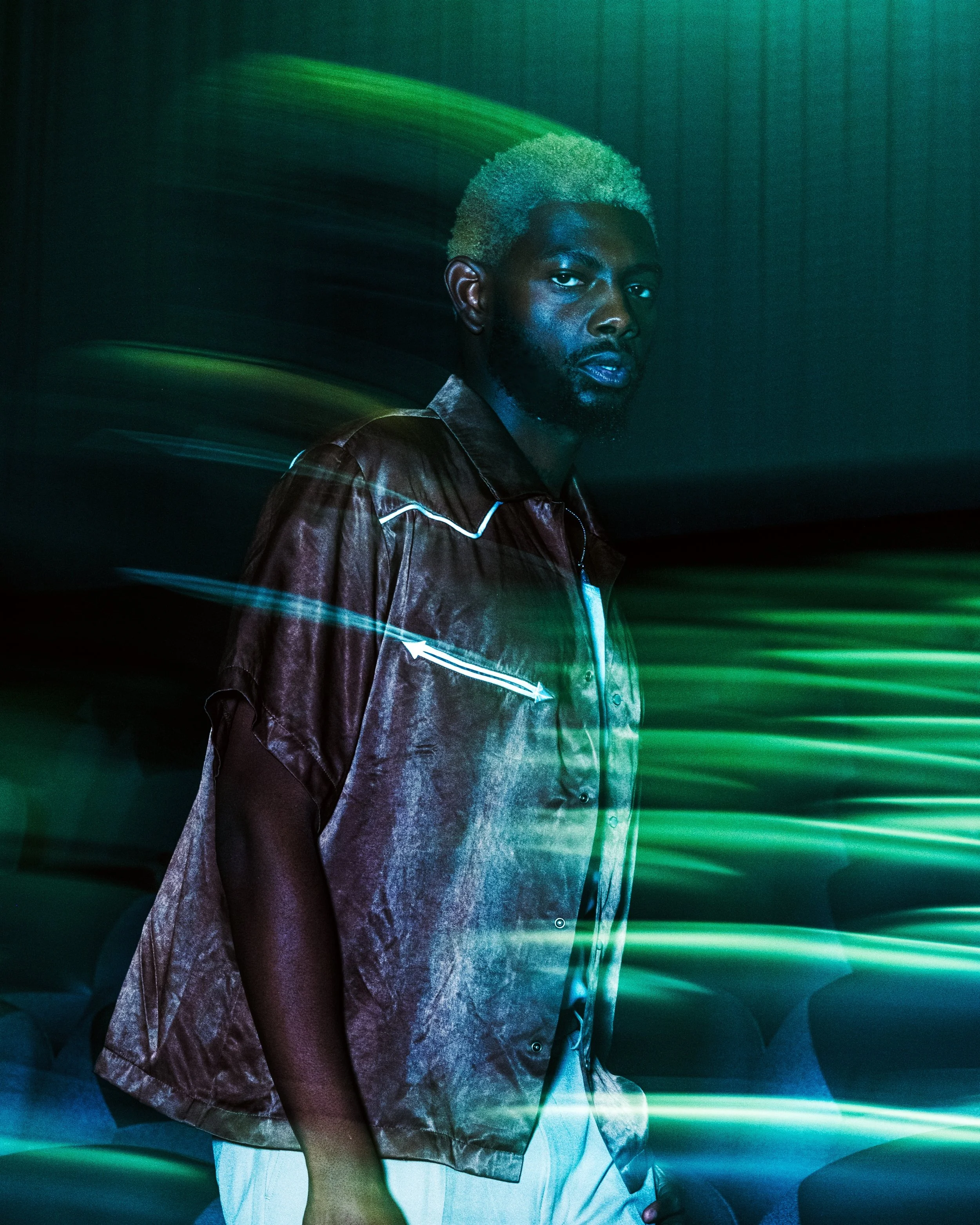 A man with bleached blonde hair and a beard stands against a dark background with green and blue light streaks.